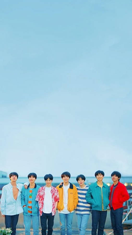 cute bts group 桌布 壁紙 — 七個穿著彩色外套的年輕男子站在一起，背景是水邊廣闊的淡藍色天空。