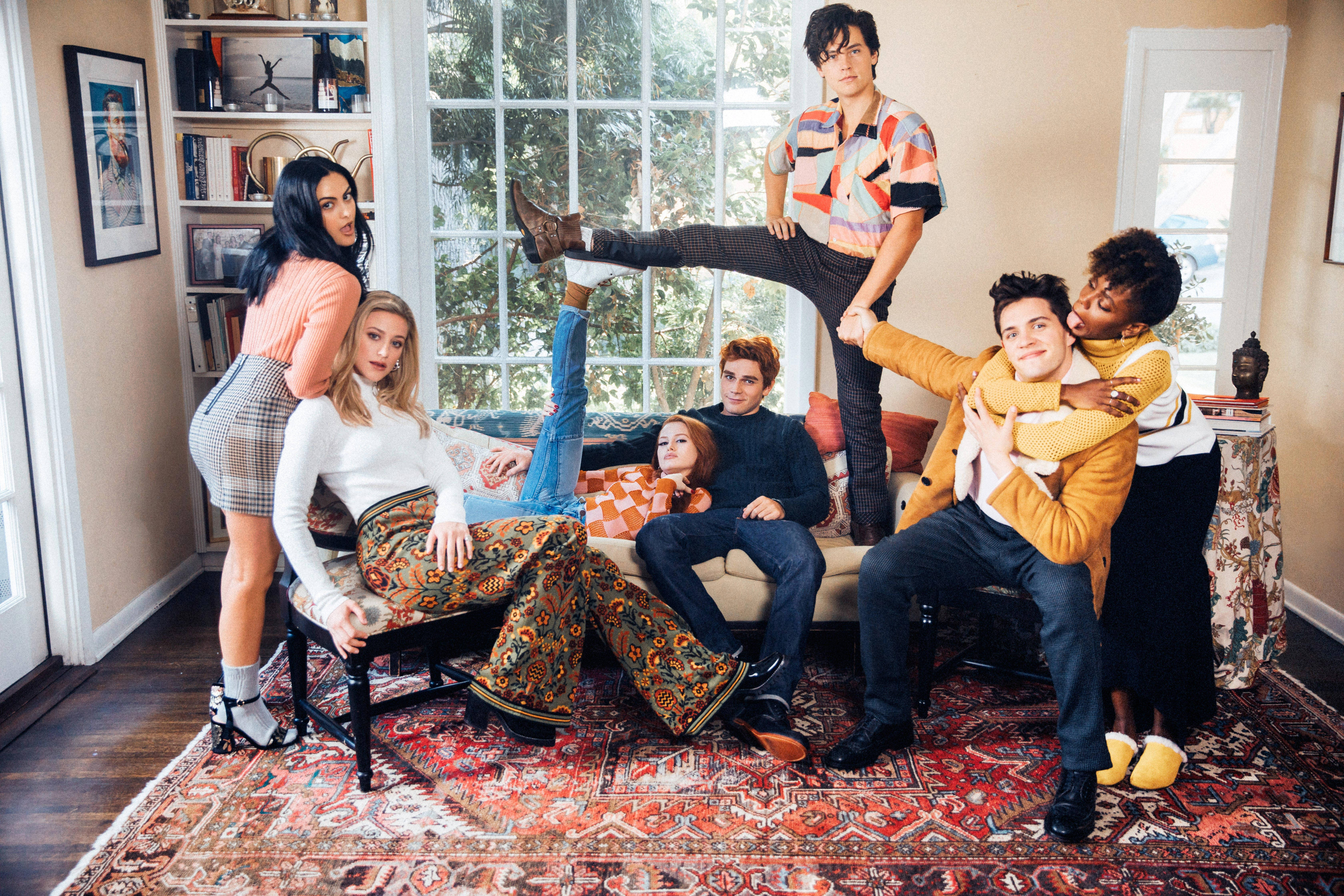 Riverdale 桌布 壁紙 — 七個穿著時尚的人在陽光充足的客廳裡擺出俏皮的姿勢，客廳裡有復古地毯和書架。