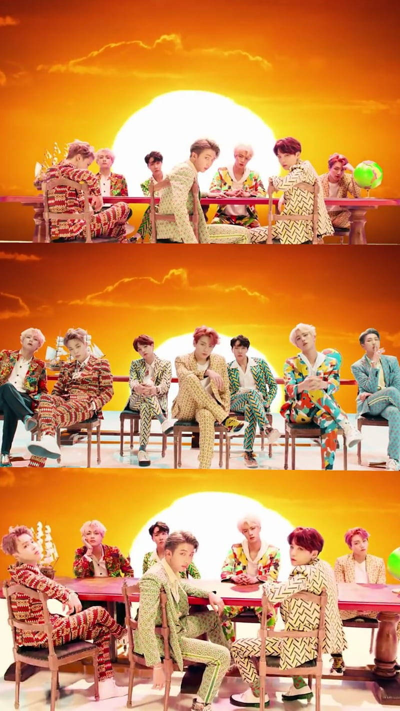 BTS 桌布 壁紙 — 七個穿著色彩繽紛圖案西裝的男人圍坐在一張紅色桌子旁，背景是充滿活力的橙色日落和一個巨大的太陽。