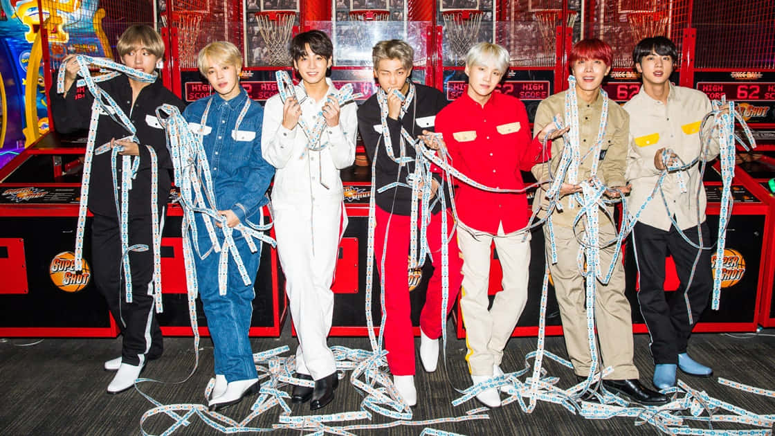 BTS 桌布 壁紙 — 七個穿著色彩繽紛服裝的男人站在遊戲機中心，拿著纏繞的軟尺，身後有遊戲機。