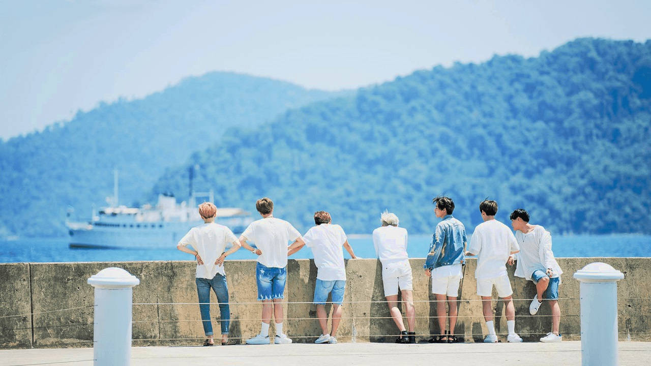 BTS 桌布 桌布 壁紙 — 七個身穿白色服裝的年輕男子背對背站在碼頭上，凝視著一艘在晴朗藍天下駛過翠綠山丘的郵輪。