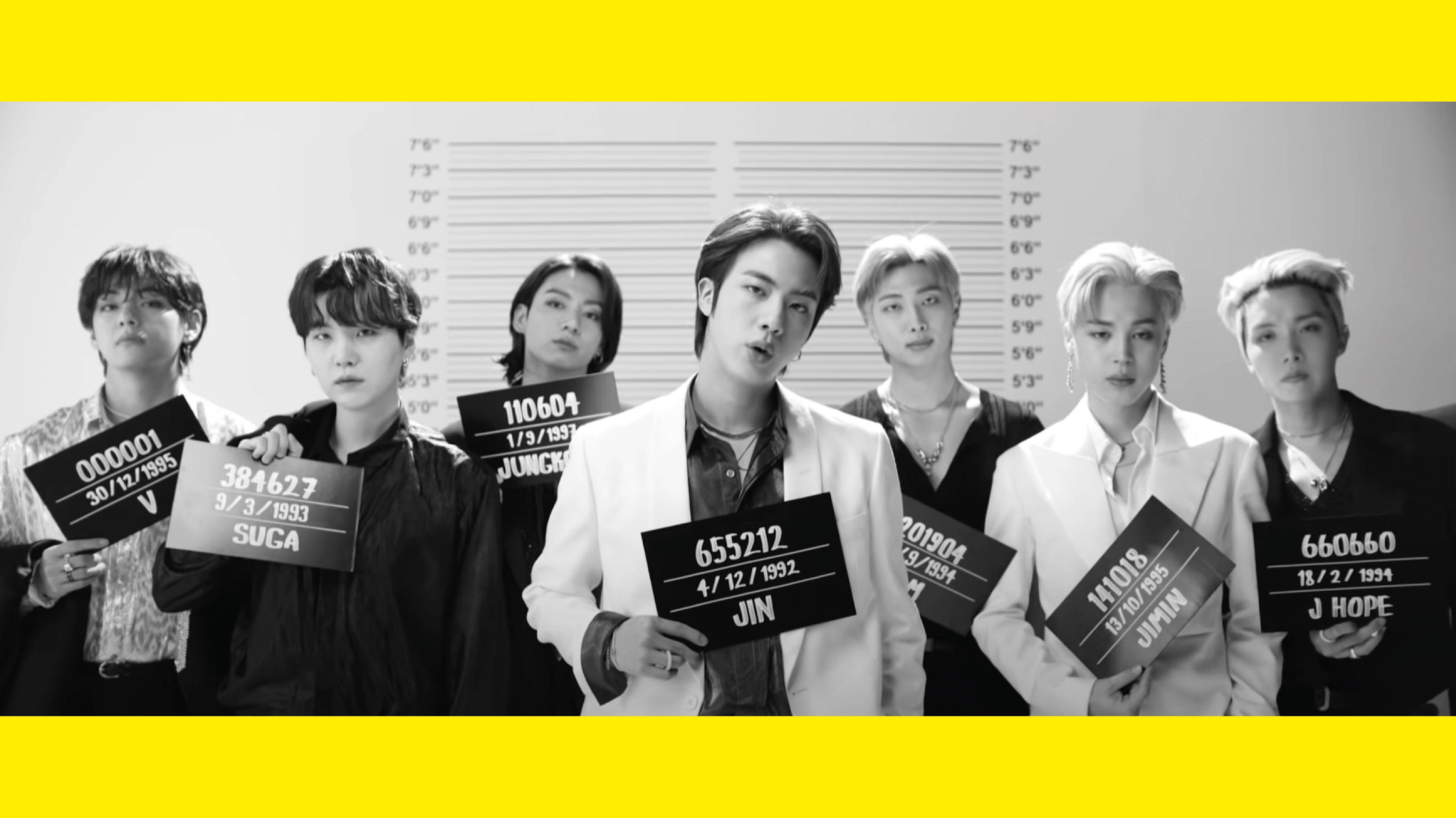 BTS 桌布 桌布 壁紙 — 七名男子以疑犯照風格排成一列，手持身高圖表和身份證，在純白背景前擺姿勢，背景由大膽的黃色邊框框住。