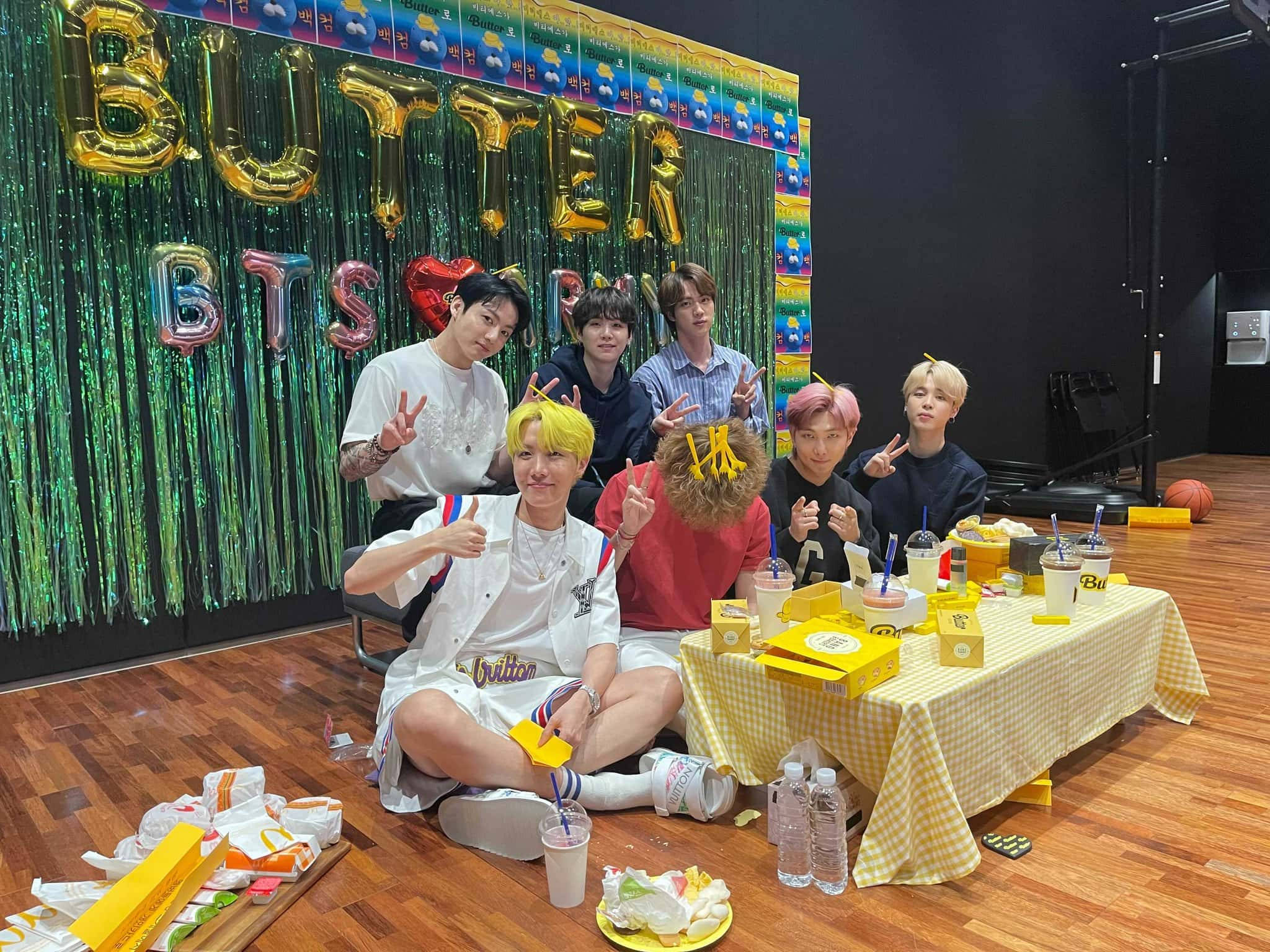 BTS 2021 桌布 壁紙 — 七名身穿休閒服裝的男子，在一個黃色格子桌布、擺滿飲料和點心的桌子旁愉快地擺姿勢，背景是閃閃發光的綠色，並有金色的「BUTTER BTS」氣球。
