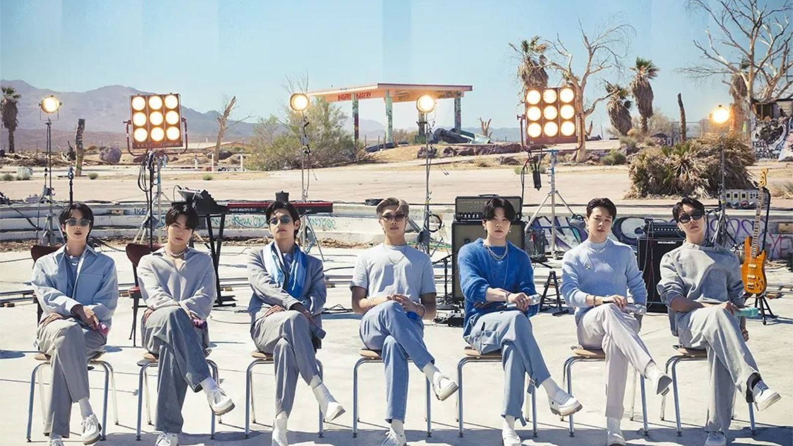 BTS 桌布 桌布 壁紙 — 七名身穿協調淺藍色服裝的男子坐在沙漠加油站場地的椅子上，在明亮的攝影棚燈光下。