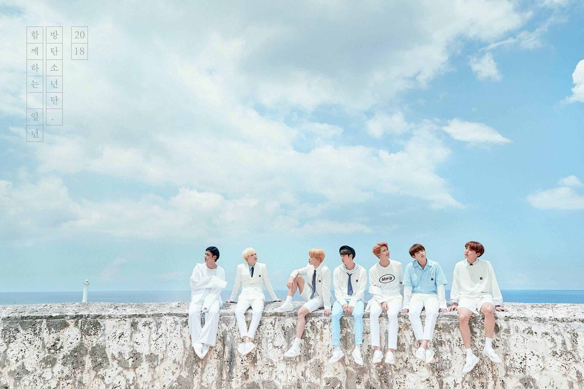 BTS 桌布 桌布 壁紙 — 七名身穿白色服裝的年輕男子隨意地坐在俯瞰海洋的風化石牆上，背景是藍天白雲。