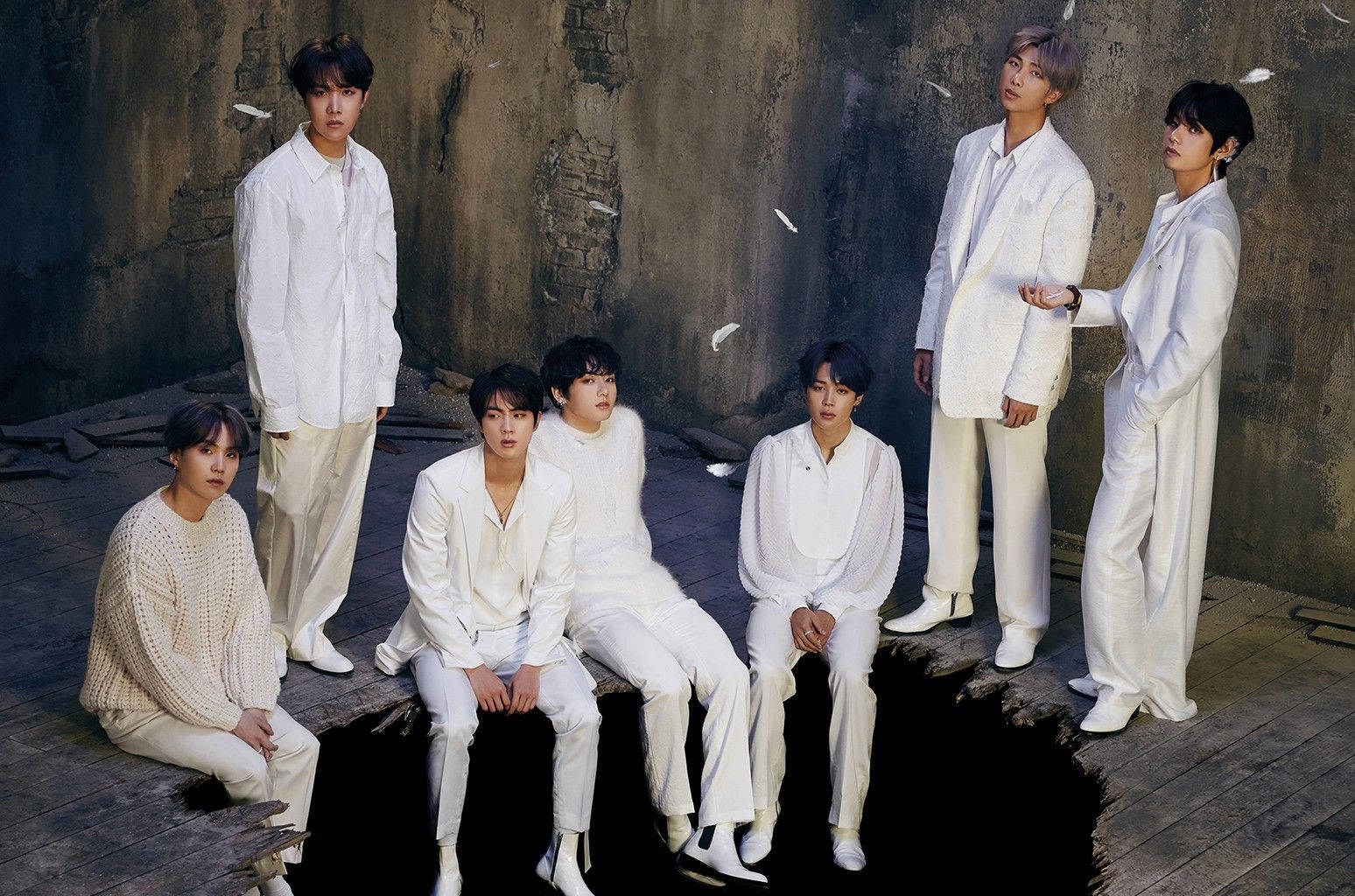 BTS 桌布 桌布 壁紙 — 七名身穿白色服裝的男子，在風化的混凝土牆前，伴隨著飄落的白色羽毛，莊嚴地擺出姿勢。