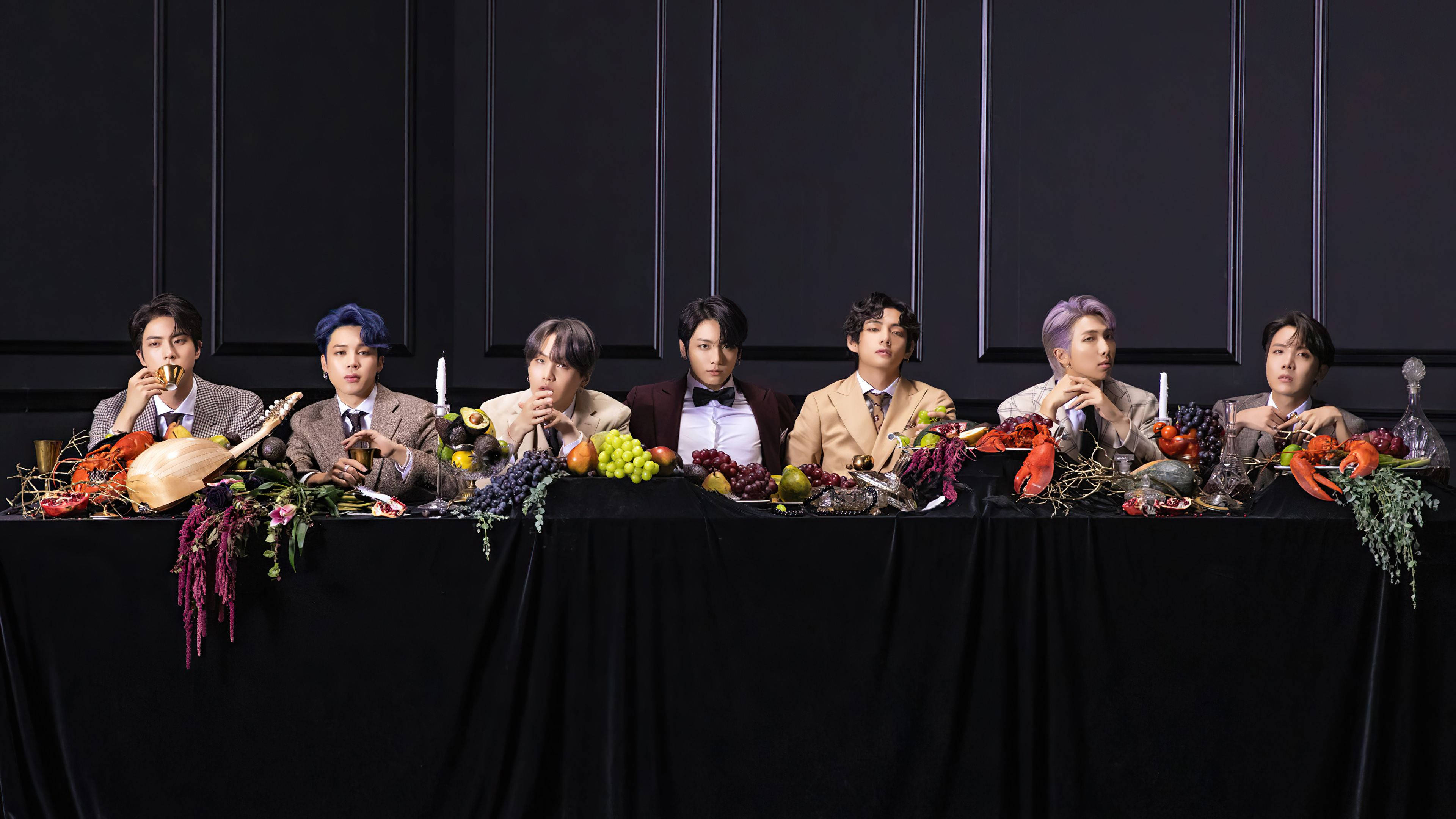 BTS 桌布 壁紙 — 七名身穿西裝的男子坐在一張鋪著黑布的桌子旁，桌上擺滿了色彩繽紛的水果和海鮮，背景是深色鑲板牆。