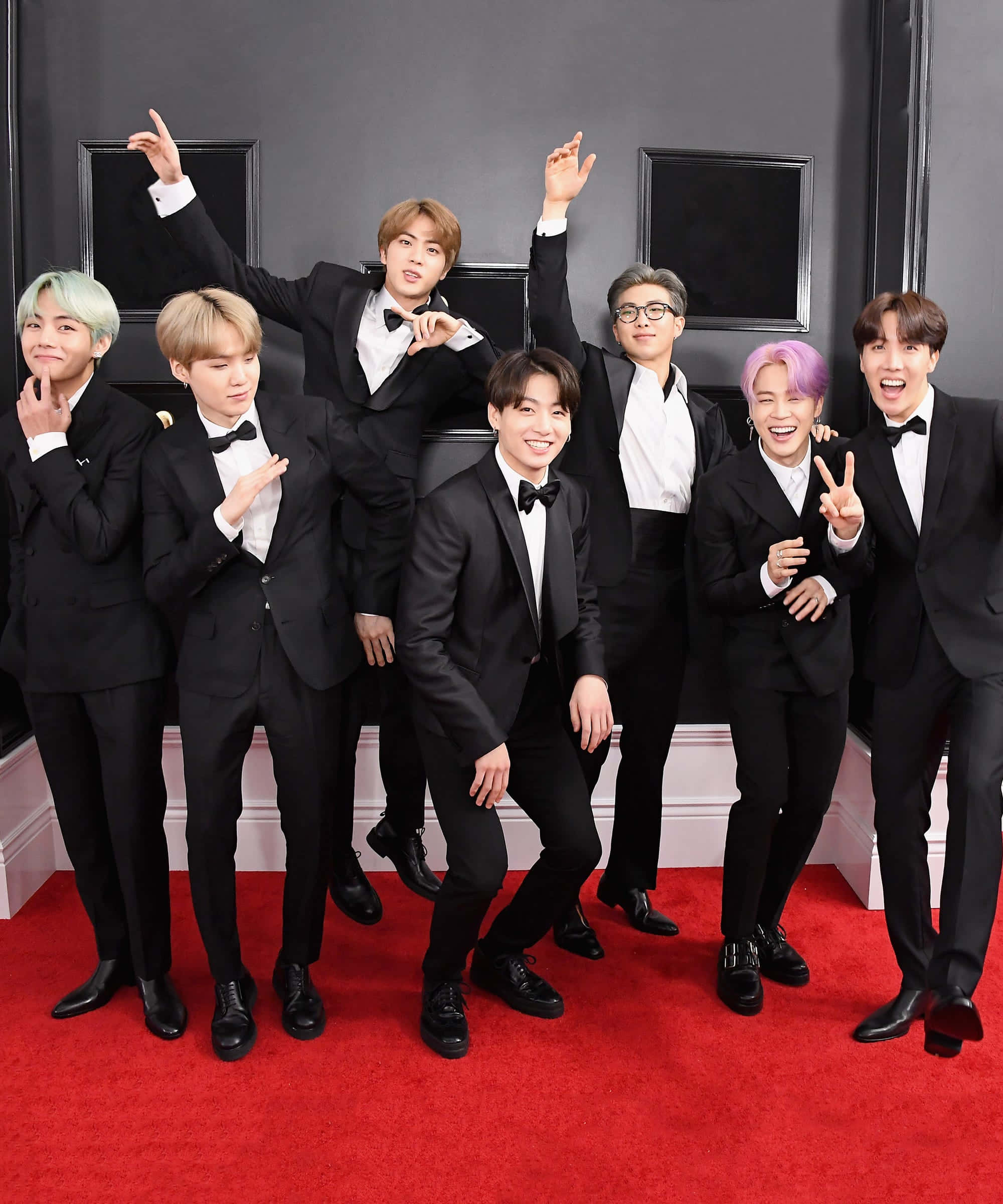 BTS 桌布 壁紙 — 七名身穿黑色燕尾服的男士在鮮紅地毯上擺出俏皮姿勢，背景是帶有框格鑲板的深灰色牆壁。