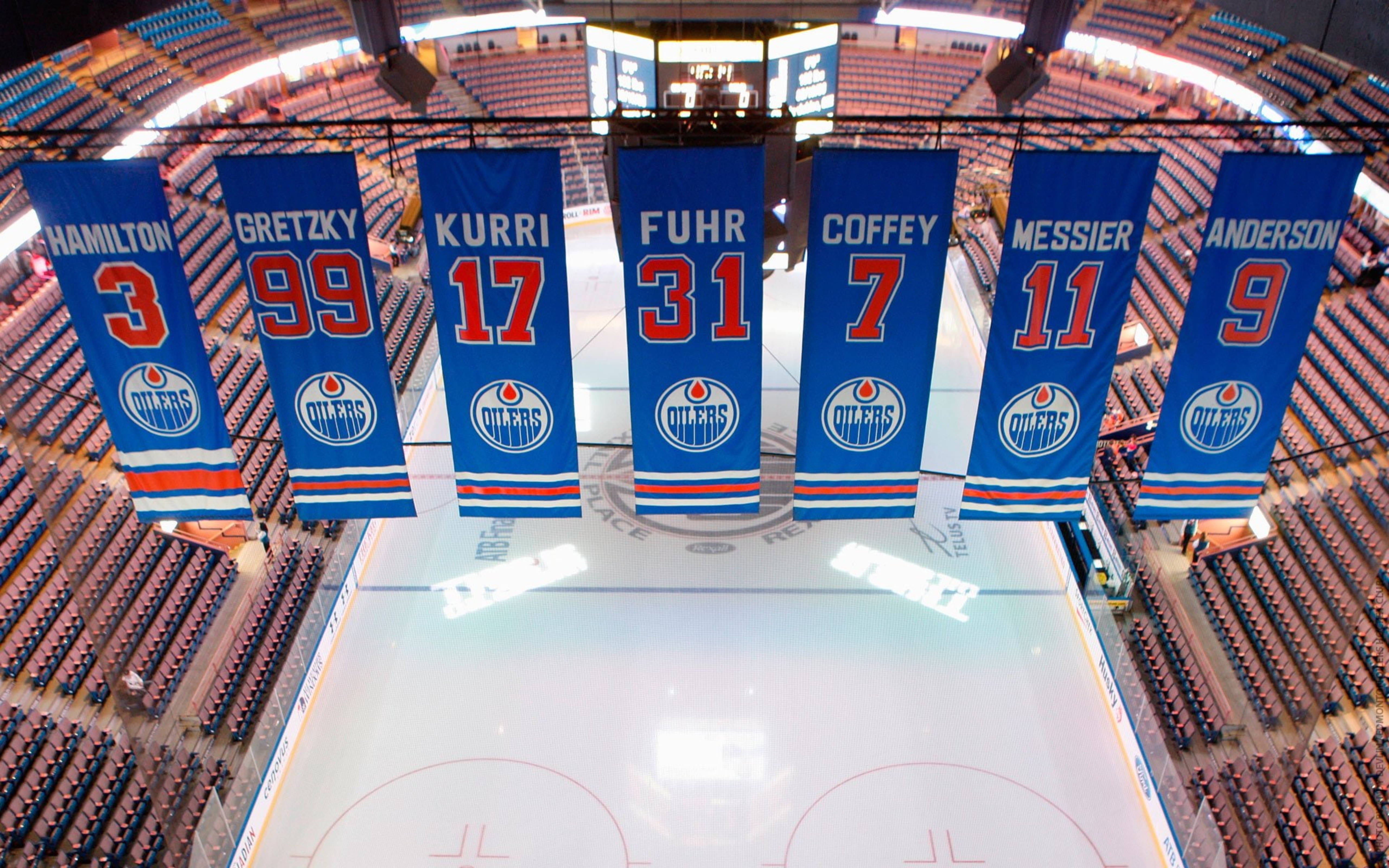 Oilers 桌布 壁紙 — 七面藍色的 Edmonton Oilers 退役球衣旗幟在一個擠滿觀眾的場館中，驕傲地懸掛在完美無瑕的冰面之上。