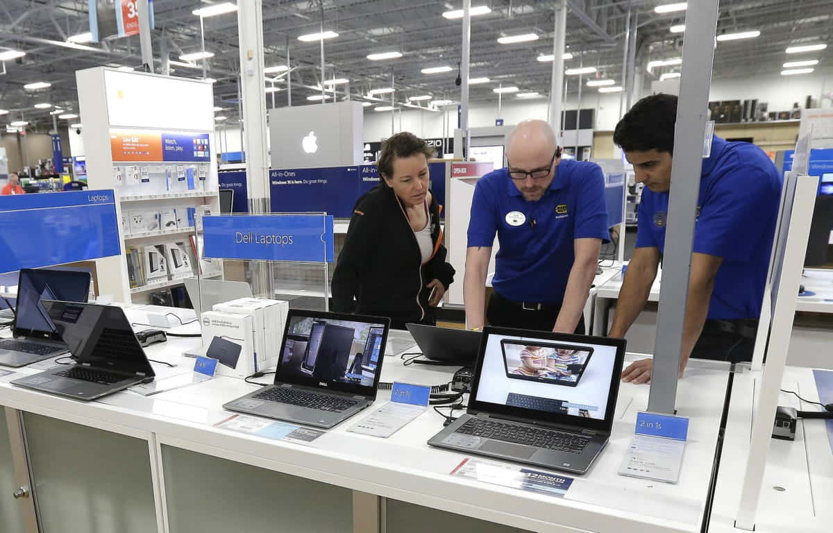 Best Buy 桌布 壁紙 — 三人在光線明亮的電子產品店內，在品牌標誌和天花燈光下查看展示中的筆記型電腦。