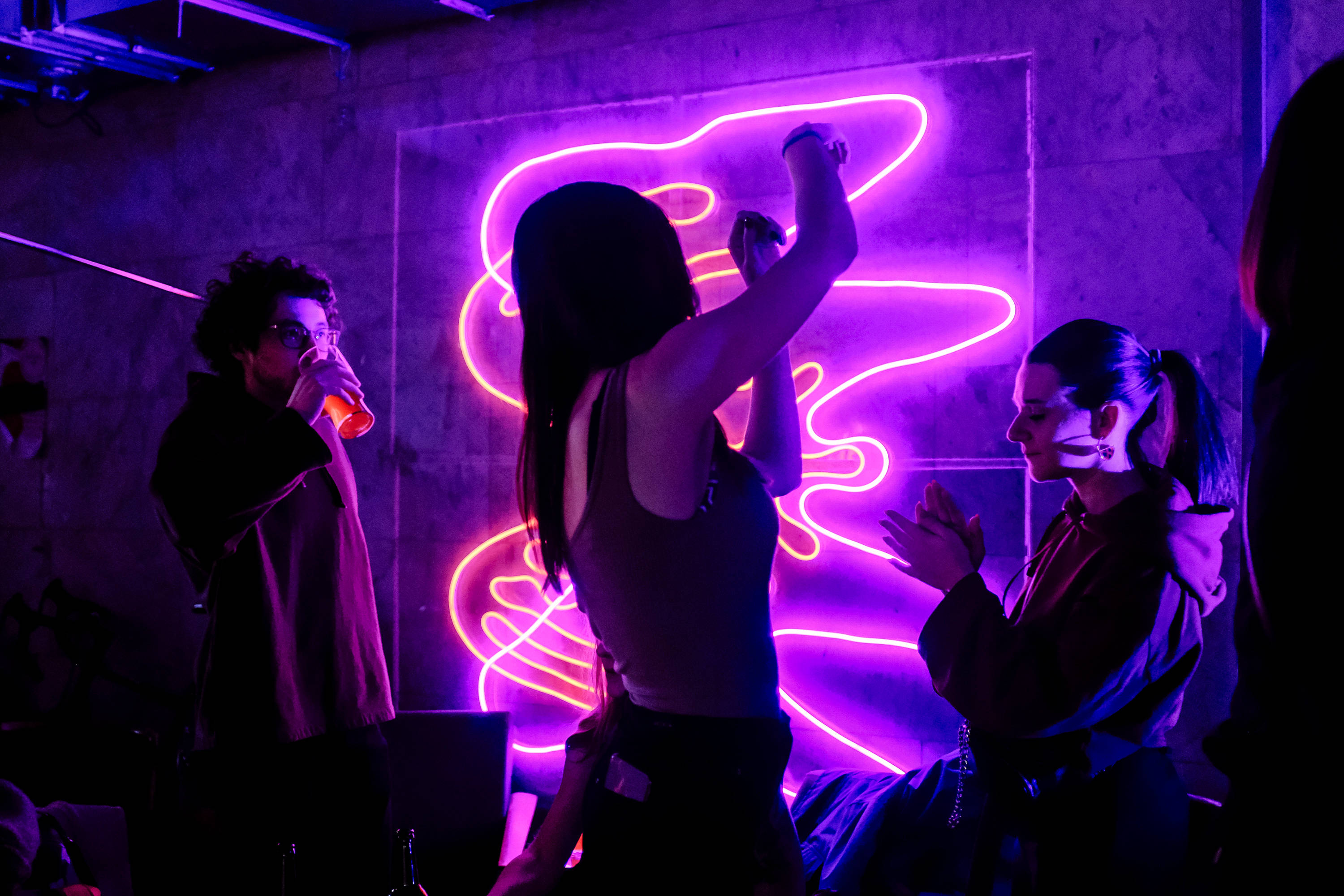 dance party 桌布 壁紙 — 三人在昏暗的房間中社交，房間被發光的牆壁藝術裝置散發出的鮮豔紫色霓虹燈光所沐浴。