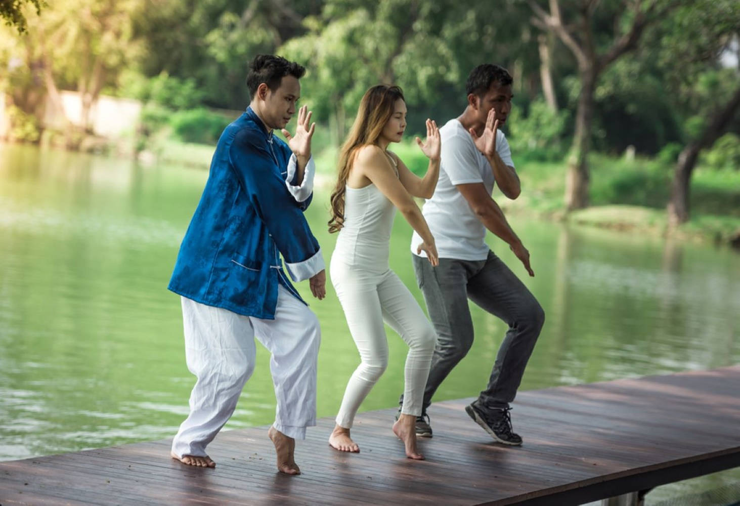 Exercise 桌布 壁紙 — 三人在木製碼頭上以同步姿勢練習武術，旁邊是平靜的綠色湖泊，周圍環繞著茂密的樹木。