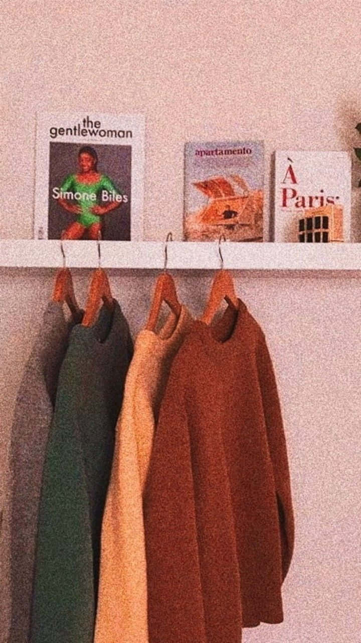 Clothes 桌布 壁紙 — 三件舒適的毛衣掛在一個展示時尚雜誌的架子下方，背景是柔和的粉紅色牆壁。