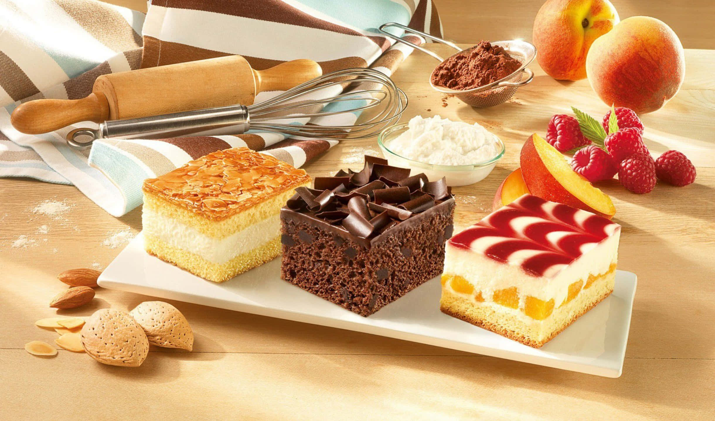 2440x1440 Desserts 桌布 壁紙 — 三件頹廢甜品切片杏仁蛋糕、巧克力蛋糕和蜜桃奶油蛋糕放在白色盤子上，搭配新鮮水果和烘焙工具在木桌上。