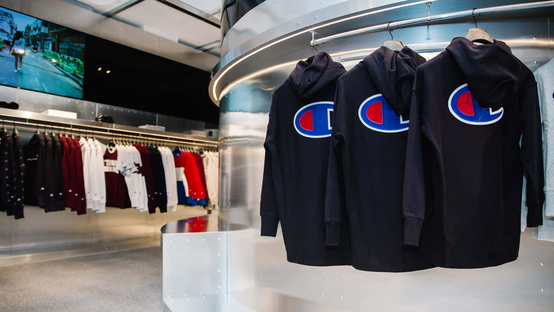 Champion 桌布 壁紙 — 三件黑色Champion連帽上衣掛在彎曲的金屬展示架上，背景是現代零售店，有衣架和大型螢幕。