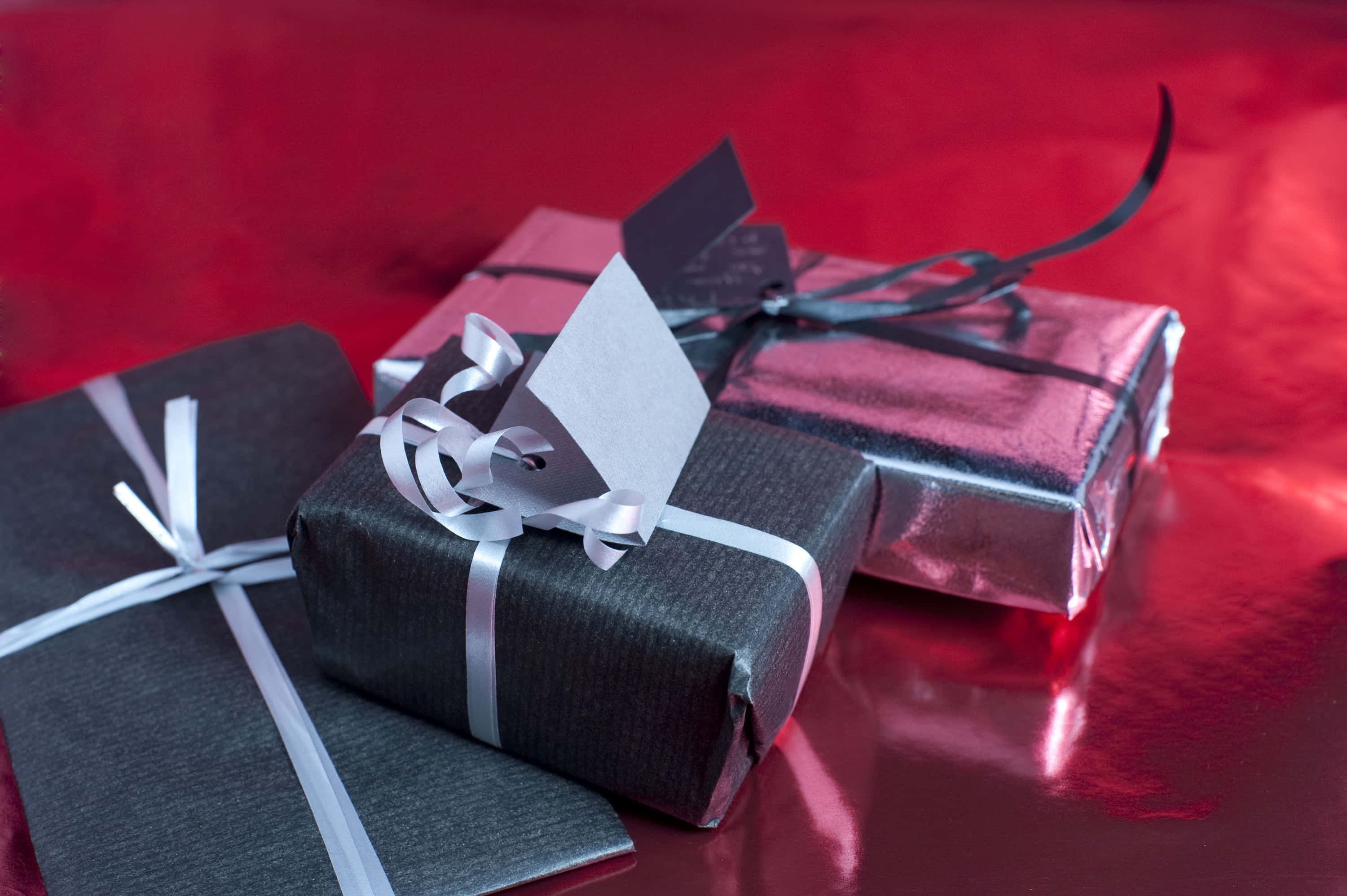 聖誕節 桌布 壁紙 — 三份以黑色和銀色包裝紙優雅包裹並繫上絲帶的禮物，放置在光滑的紅色表面上。