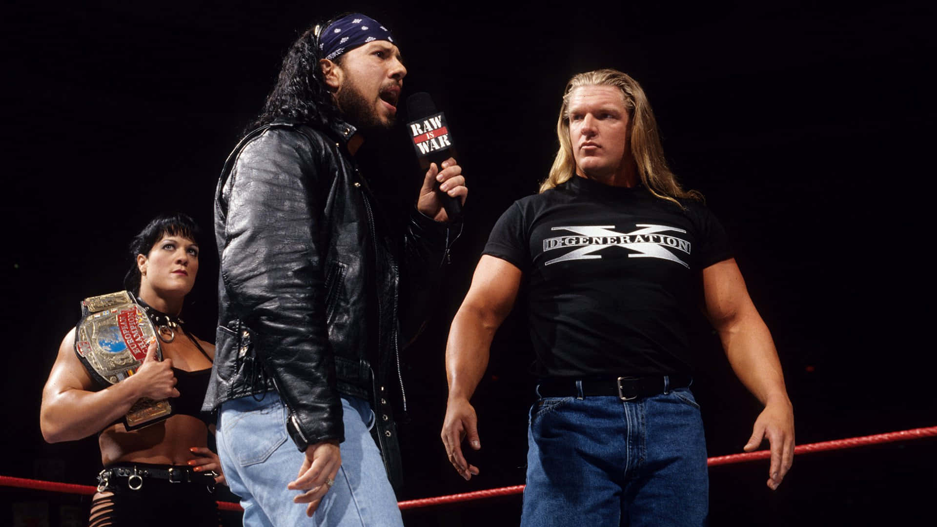 shawn michaels 桌布 壁紙 — 三位WWE摔角手站在擂台上，一人手持麥克風，另一人持有冠軍腰帶，在戲劇性的舞台燈光下。