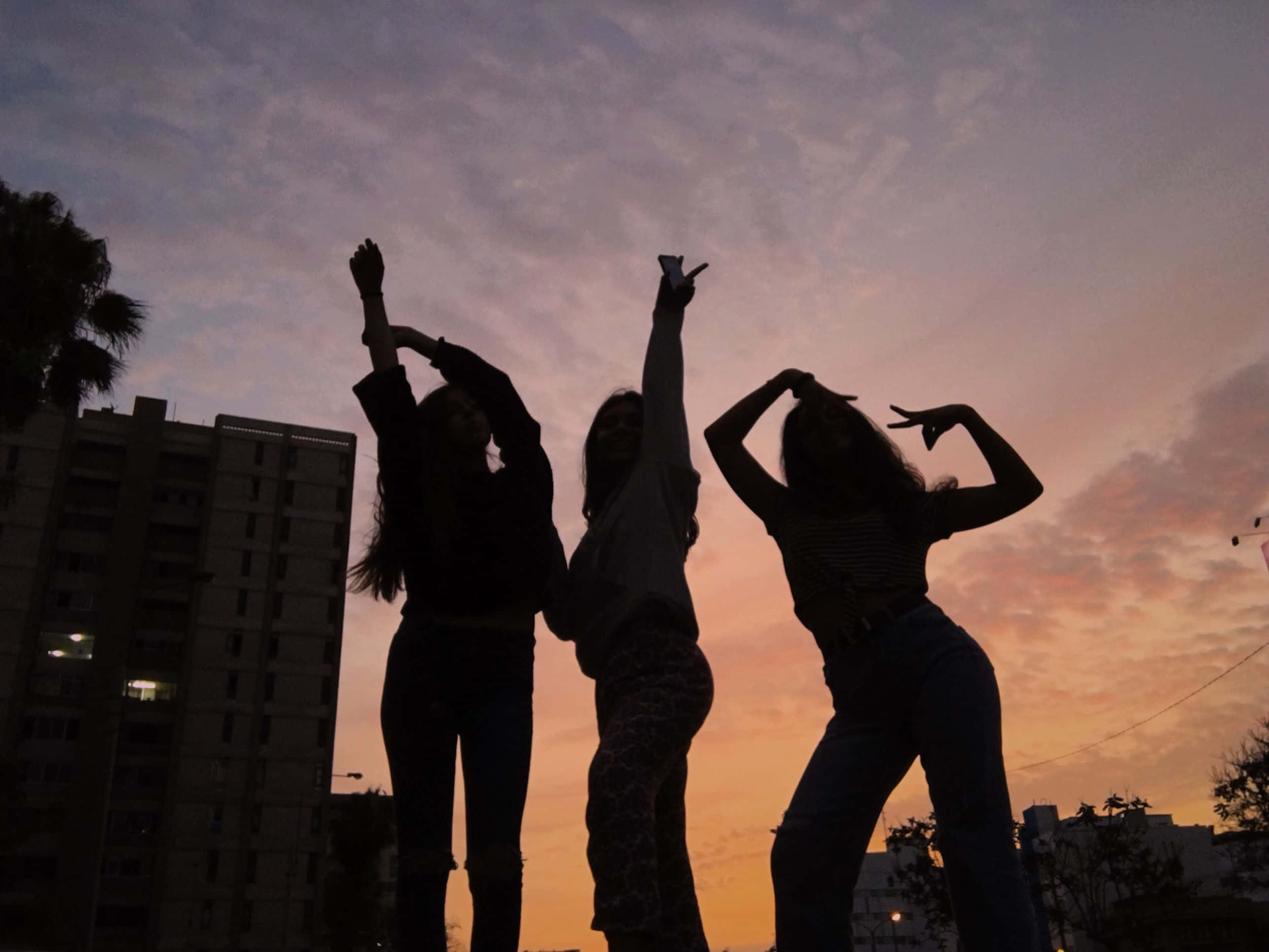 Friends 桌布 壁紙 — 三位剪影朋友在充滿活力的日落天空背景下，以城市建築為背景擺出俏皮的舞蹈姿勢。