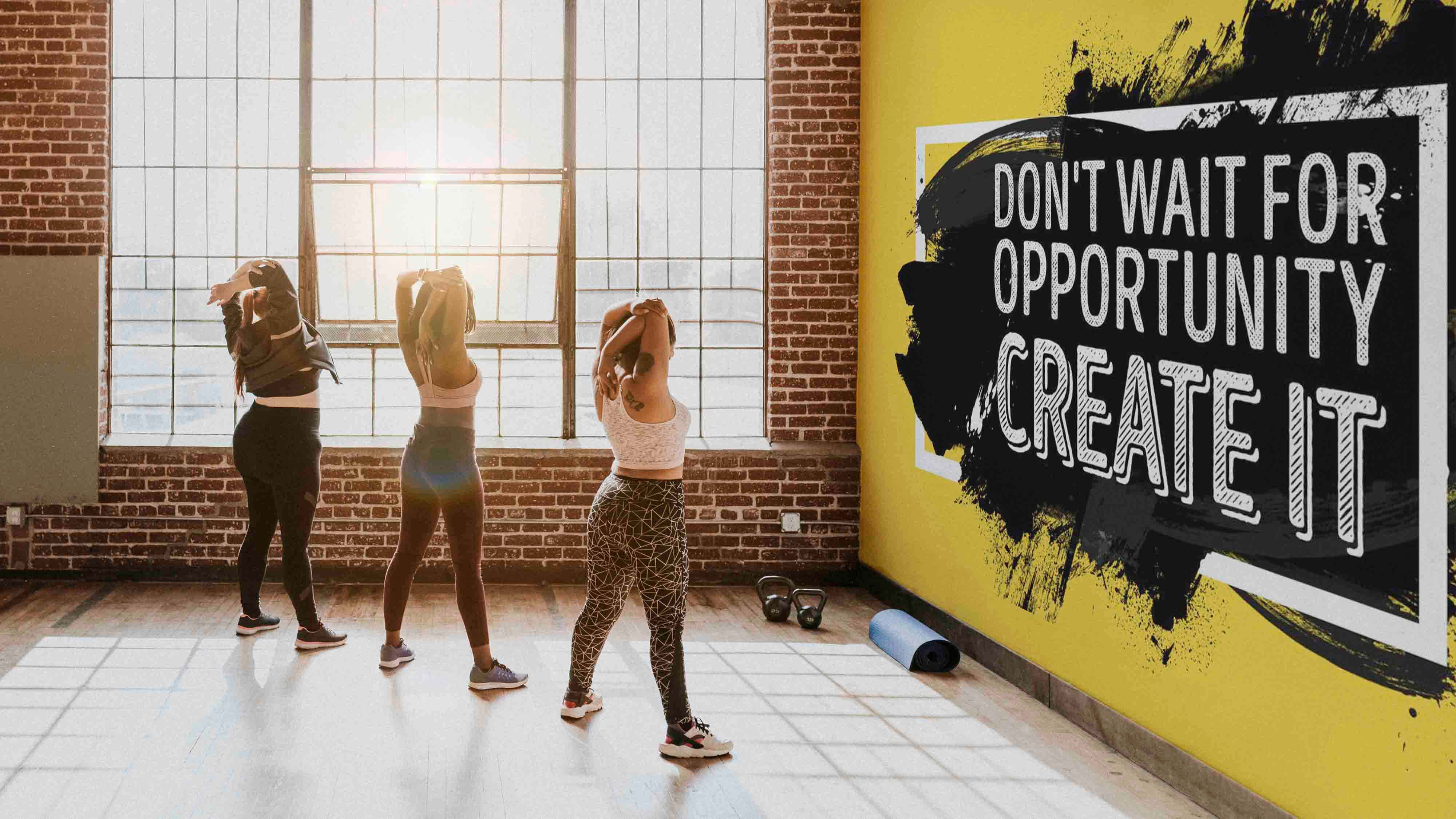 Fitness 桌布 壁紙 — 三位女士在陽光普照的磚牆健身房中伸展，黃色牆壁上印有勵志語句「DON'T WAIT FOR OPPORTUNITY CREATE IT」。
