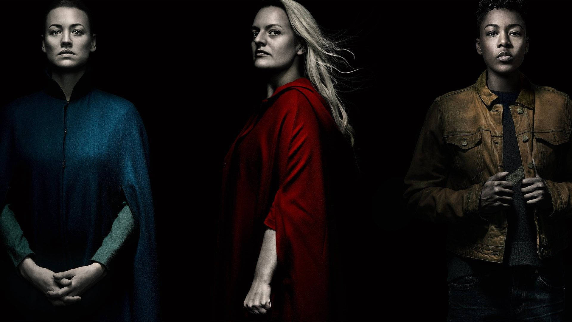 The Handmaid's Tale 桌布 壁紙 — 三位女性在昏暗的攝影棚燈光下，穿著不同顏色的服裝，莊嚴地站在黑色背景前。