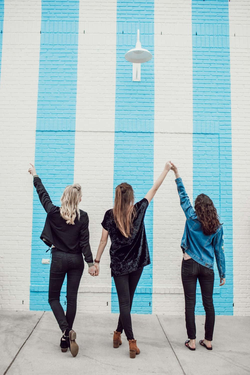 Friendship 桌布 壁紙 — 三位女性手牽手，向著街燈舉起手臂，背景是大膽的藍白條紋磚牆。