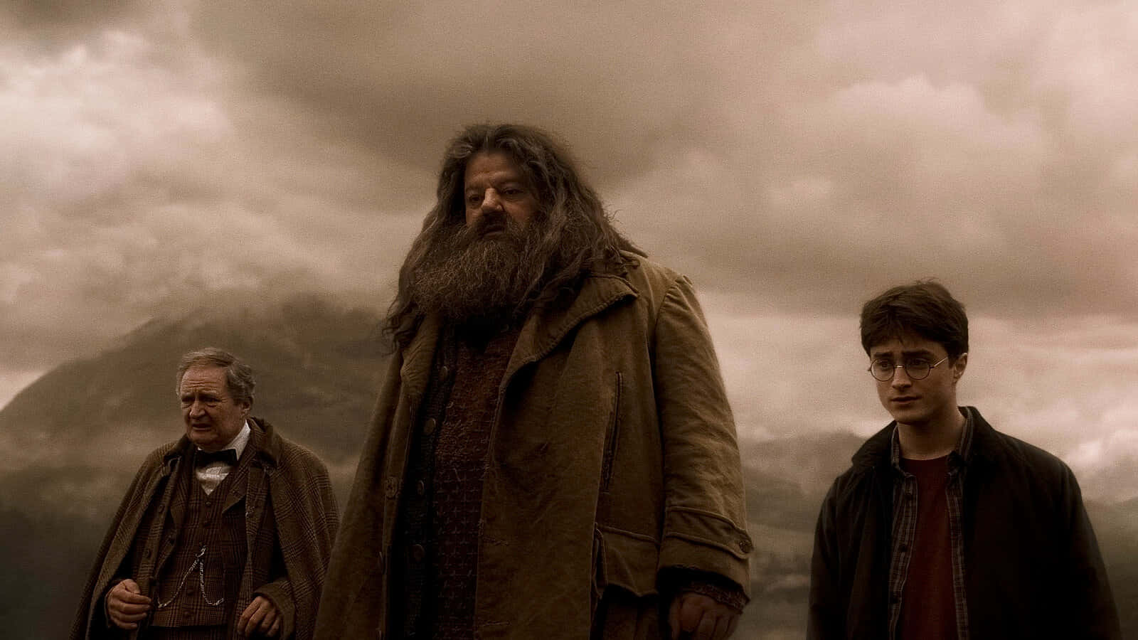 The Hogwarts Professors 桌布 壁紙 — 三位巫師在厚重的雲層下，莊嚴地站在暴風雨的山景前。