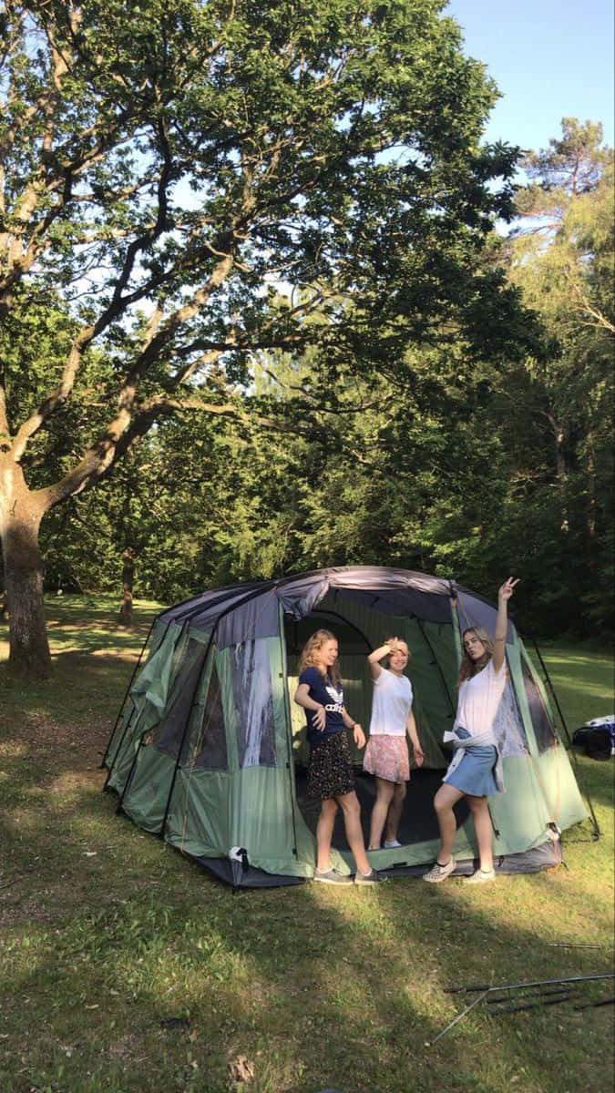 Minimalist Camping 桌布 壁紙 — 三位年輕女性在陽光明媚的森林空地上，於枝葉茂密的樹木下，在一個綠色和灰色的露營帳篷前愉快地擺姿勢。