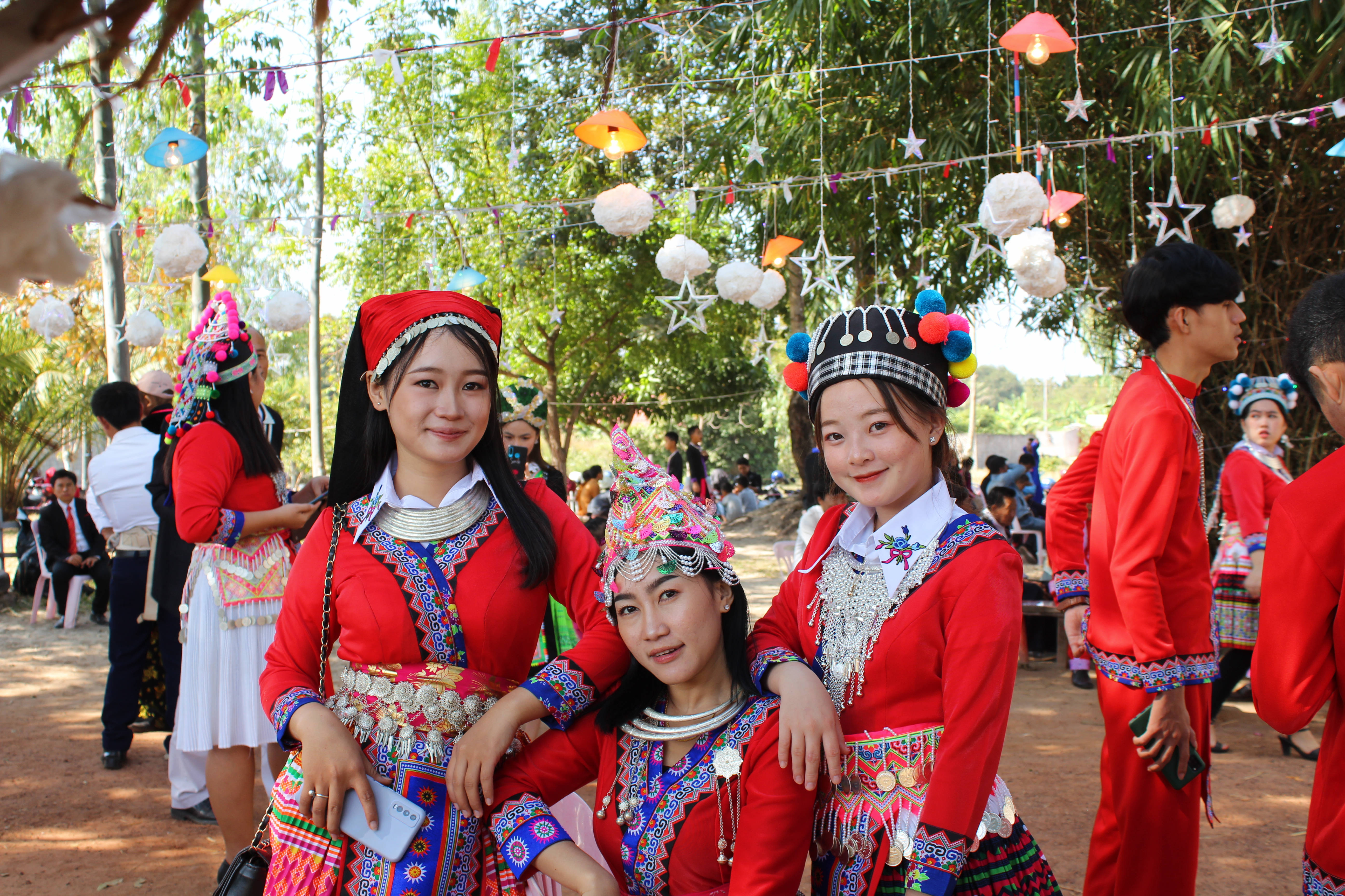 Laos 桌布 壁紙 — 三位年輕女性身穿鮮豔的傳統紅色服飾，佩戴精緻的銀飾和色彩繽紛的頭飾，在陽光明媚的戶外聚會中，於節日燈光下溫暖地微笑著看向鏡頭。