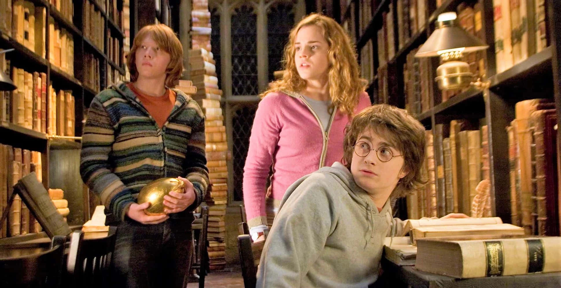 The Hogwarts Library 桌布 壁紙 — 三位年輕巫師在宏偉的圖書館中手持金色飛賊，驚訝地向上看。