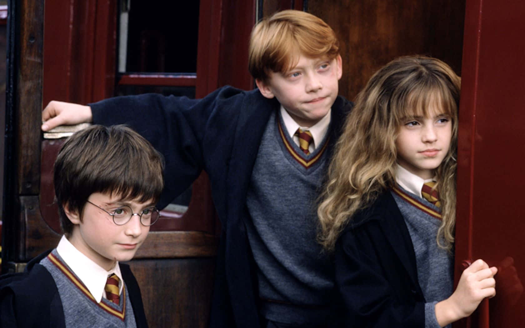 Ron Weasley 桌布 壁紙 — 三位年輕巫師穿著霍格華茲制服，神情嚴肅地站在紅色火車門旁。
