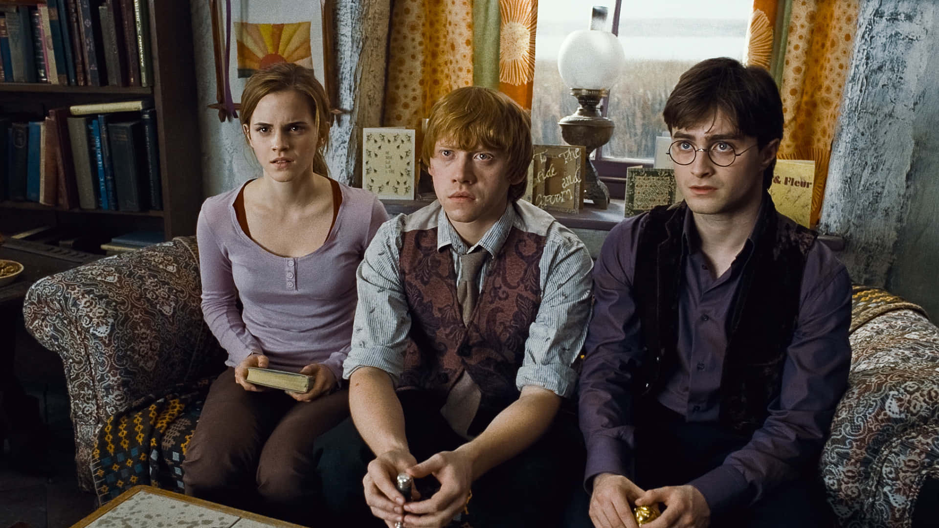 Hermione Granger 桌布 壁紙 — 三位年輕巫師莊嚴地坐在一張有圖案的沙發上，房間凌亂，堆滿書籍，燈光溫暖。
