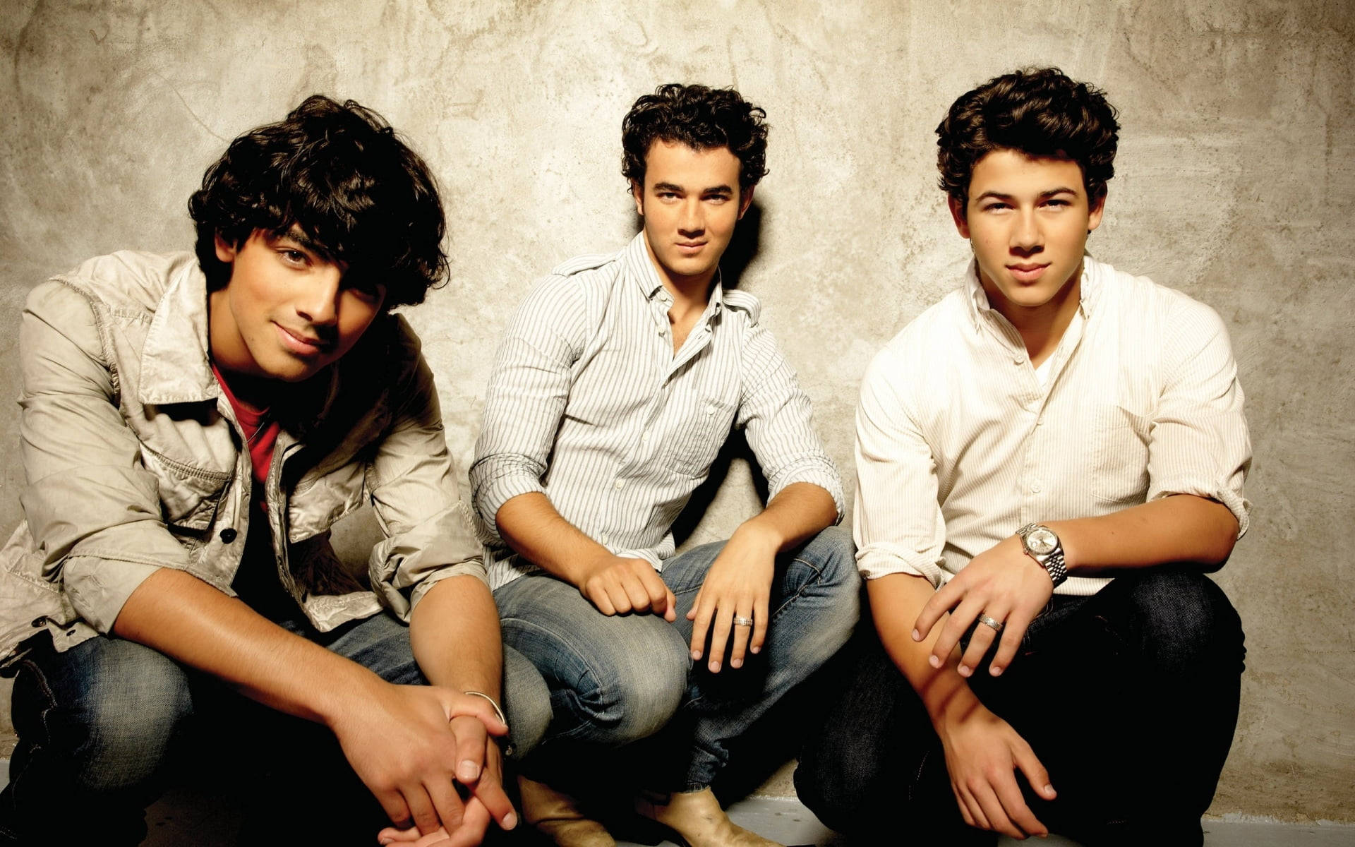 Jonas Brothers 桌布 壁紙 — 三位年輕男士身穿休閒服裝，在柔和的攝影棚燈光下，自信地以紋理灰色牆壁為背景擺姿勢。