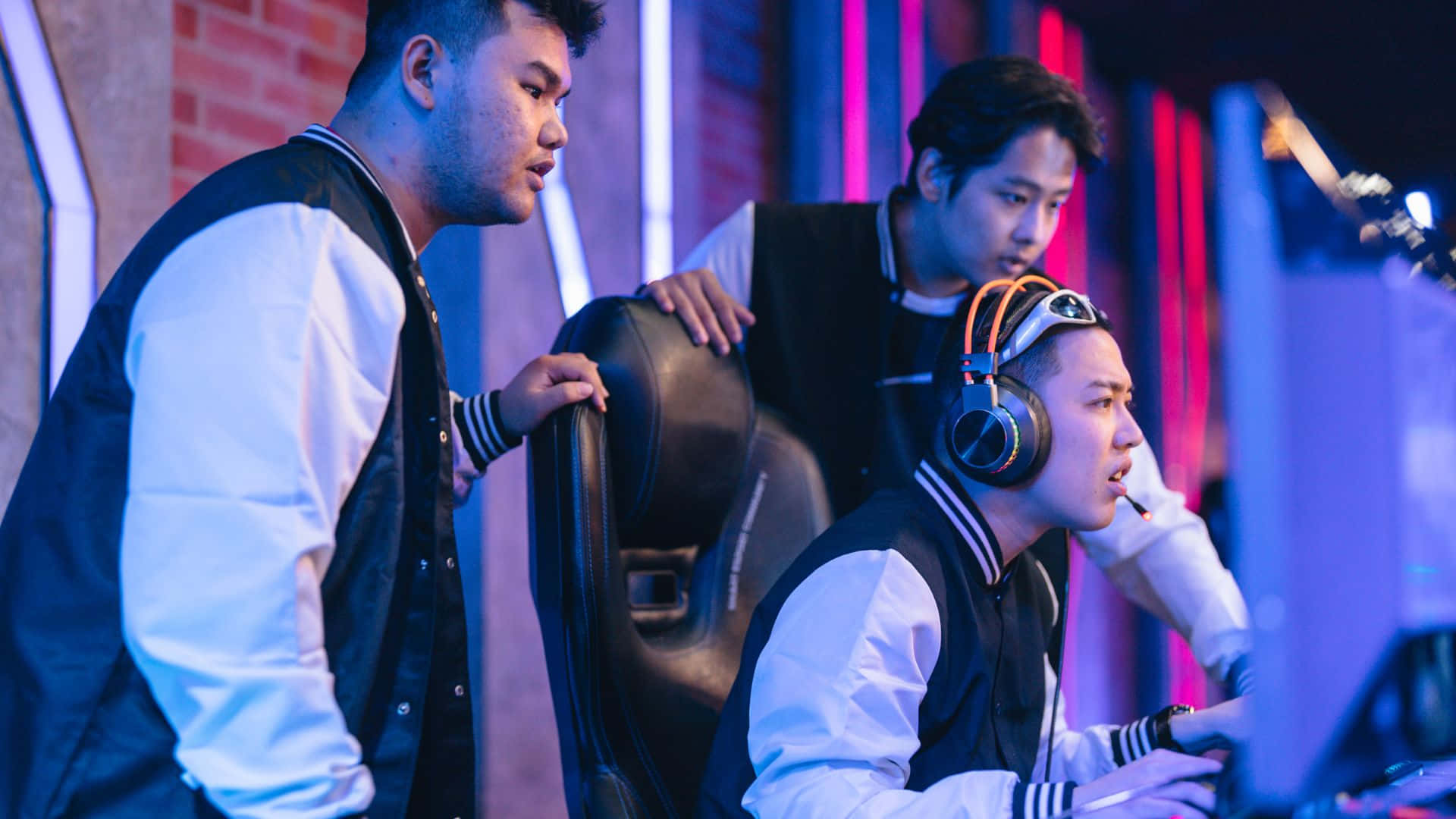 Gaming Tournaments 桌布 壁紙 — 三位年輕男子在霓虹燈照亮的遊戲室中，專注於電腦螢幕，房間有磚牆和發光的燈光。