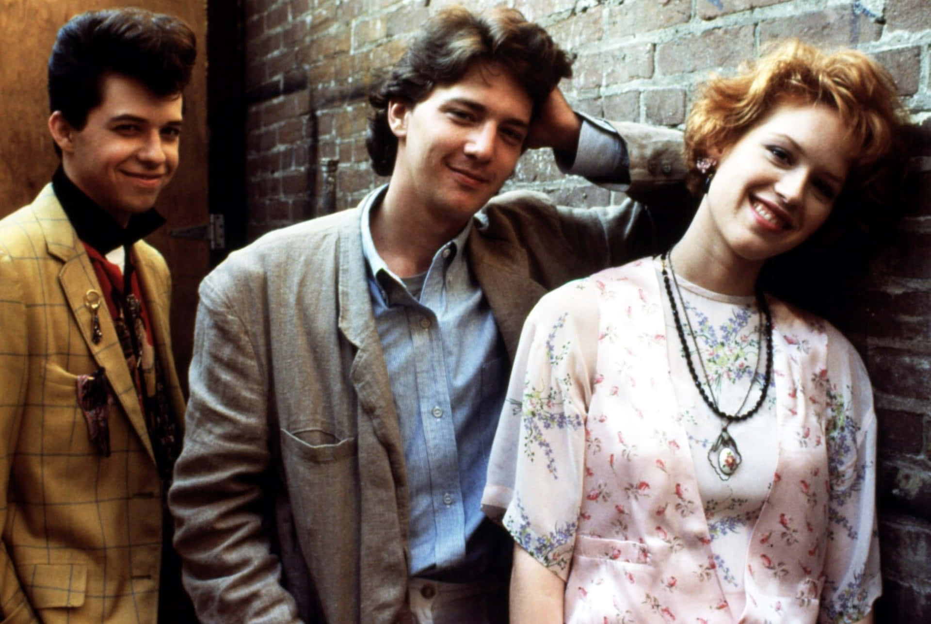 Jon Cryer 桌布 壁紙 — 三位微笑的1980年代年輕人倚靠磚牆，穿著復古格紋西裝和碎花上衣，充滿青春活力。