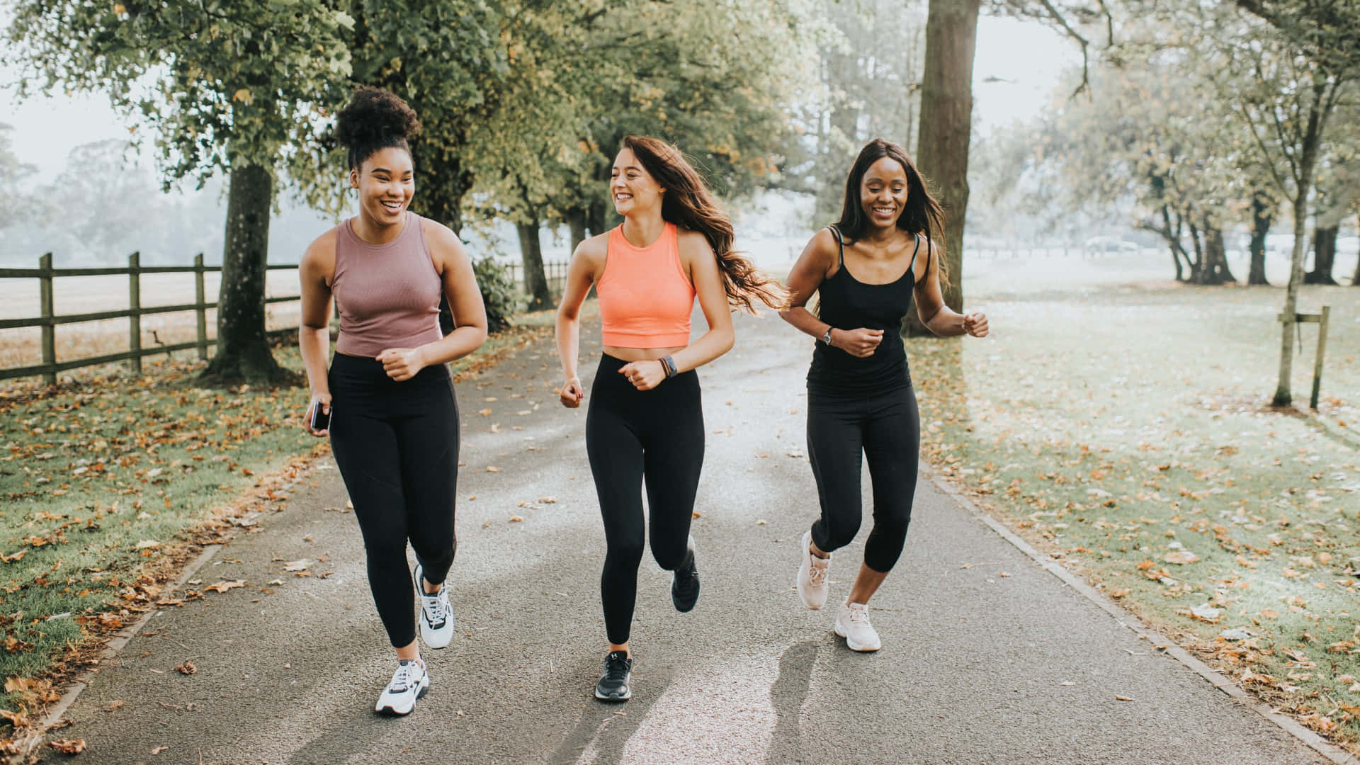 Running 桌布 壁紙 — 三位微笑的女性沿著一條兩旁種滿樹木、鋪滿秋天落葉的小徑慢跑。