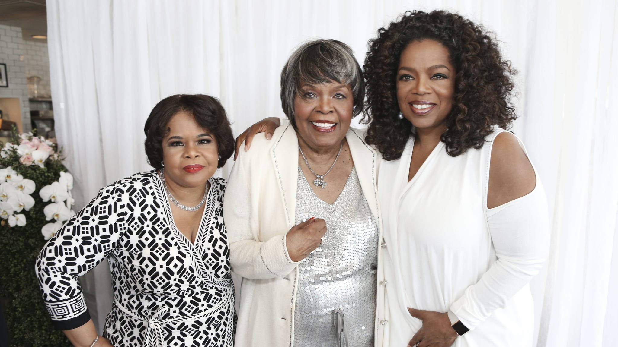 Oprah 桌布 壁紙 — 三位微笑的女性穿著優雅的服裝，在白色窗簾背景前與花卉佈置一起合影。