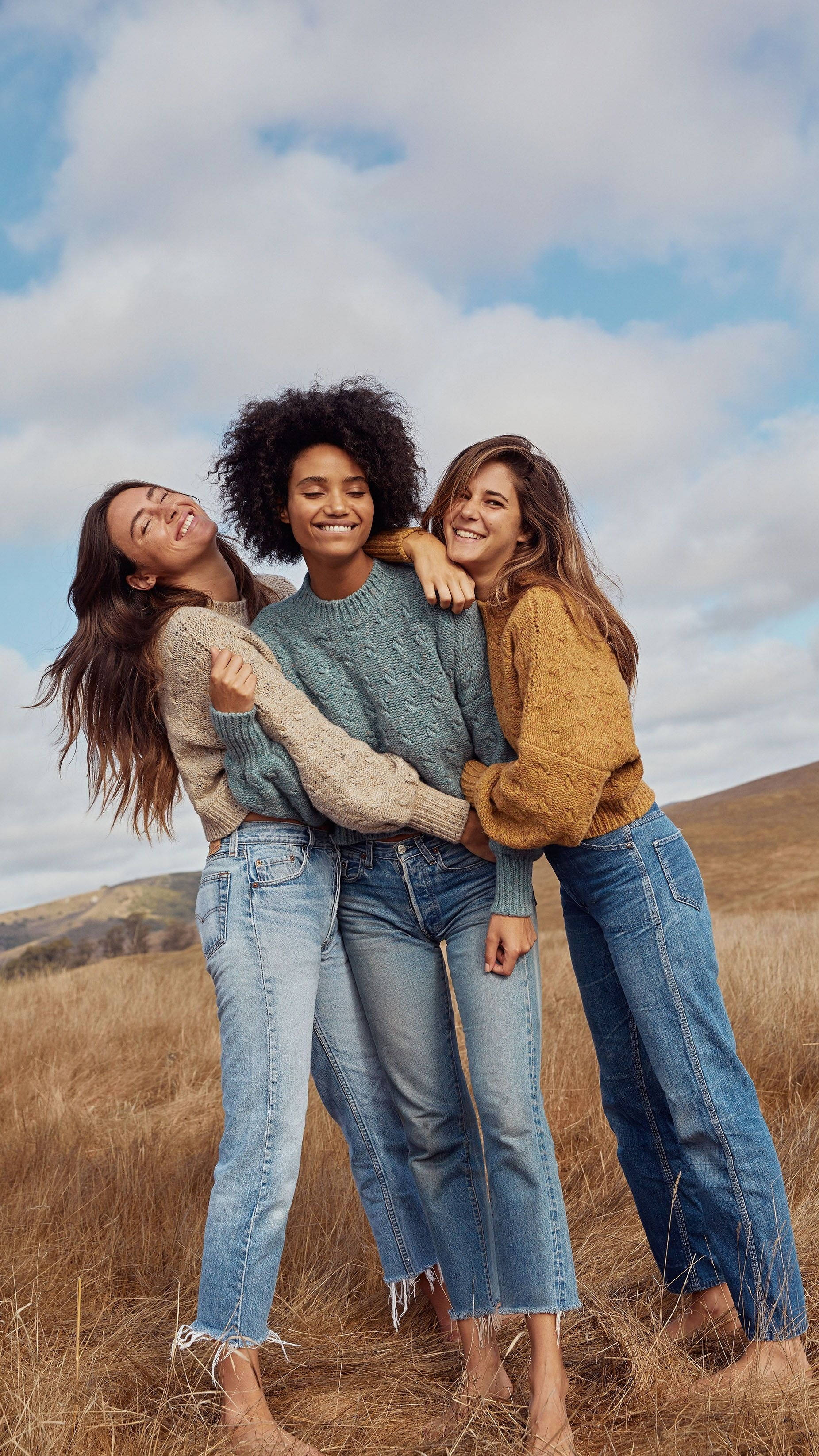 3 Best Friends 桌布 壁紙 — 三位微笑的女性穿著舒適的毛衣和牛仔褲，在部分多雲的天空下，在金色的草地上快樂地擁抱。