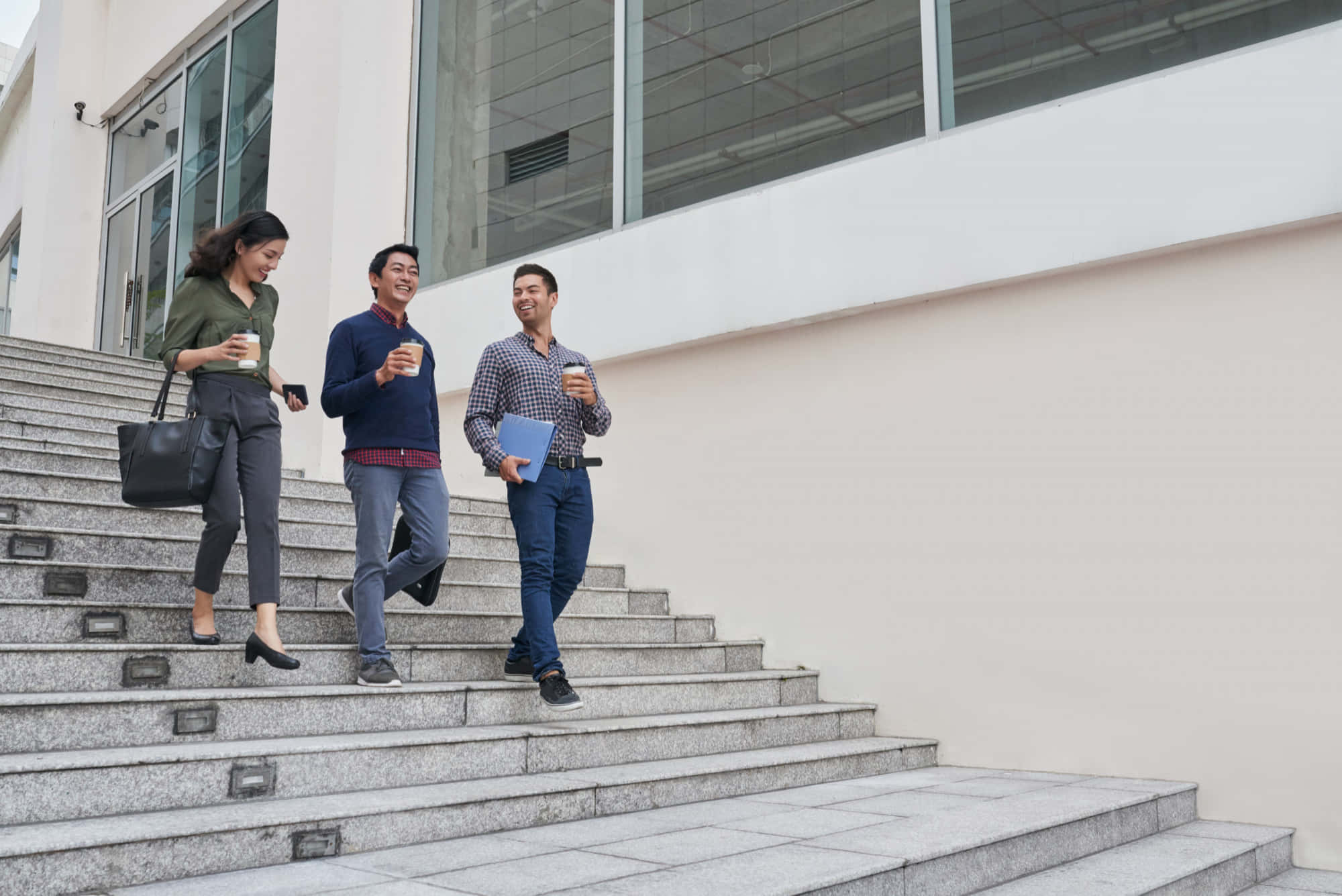 Employee 桌布 壁紙 — 三位微笑的專業人士在玻璃幕牆辦公大樓外，手持咖啡和文件，走上現代石階。