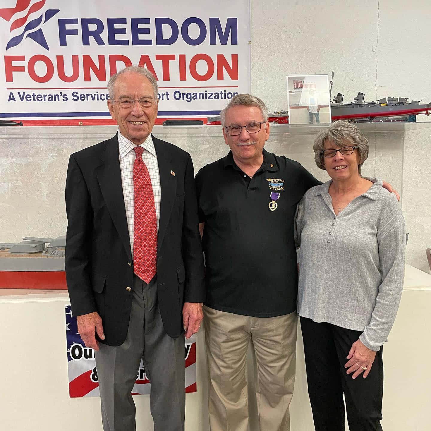 政治 桌布 壁紙 — 三位微笑的成年人一起在Freedom Foundation橫幅前合影，身後展示著模型船。