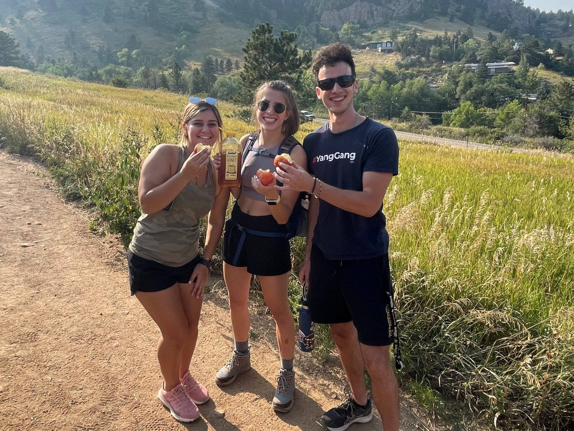 University Of Colorado At Boulder 桌布 壁紙 — 三位微笑的朋友在陽光明媚的泥土小徑上享用點心，周圍環繞著高高的草叢和遠山。