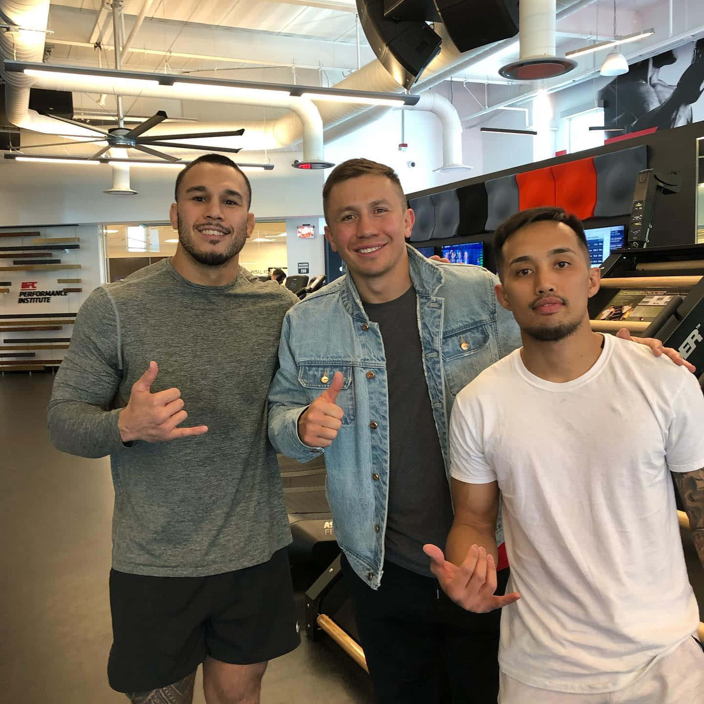 健身房 桌布 壁紙 — 三位微笑的男士在一個現代化的健身房中一起擺姿勢，可見設備和暴露的天花板管道。