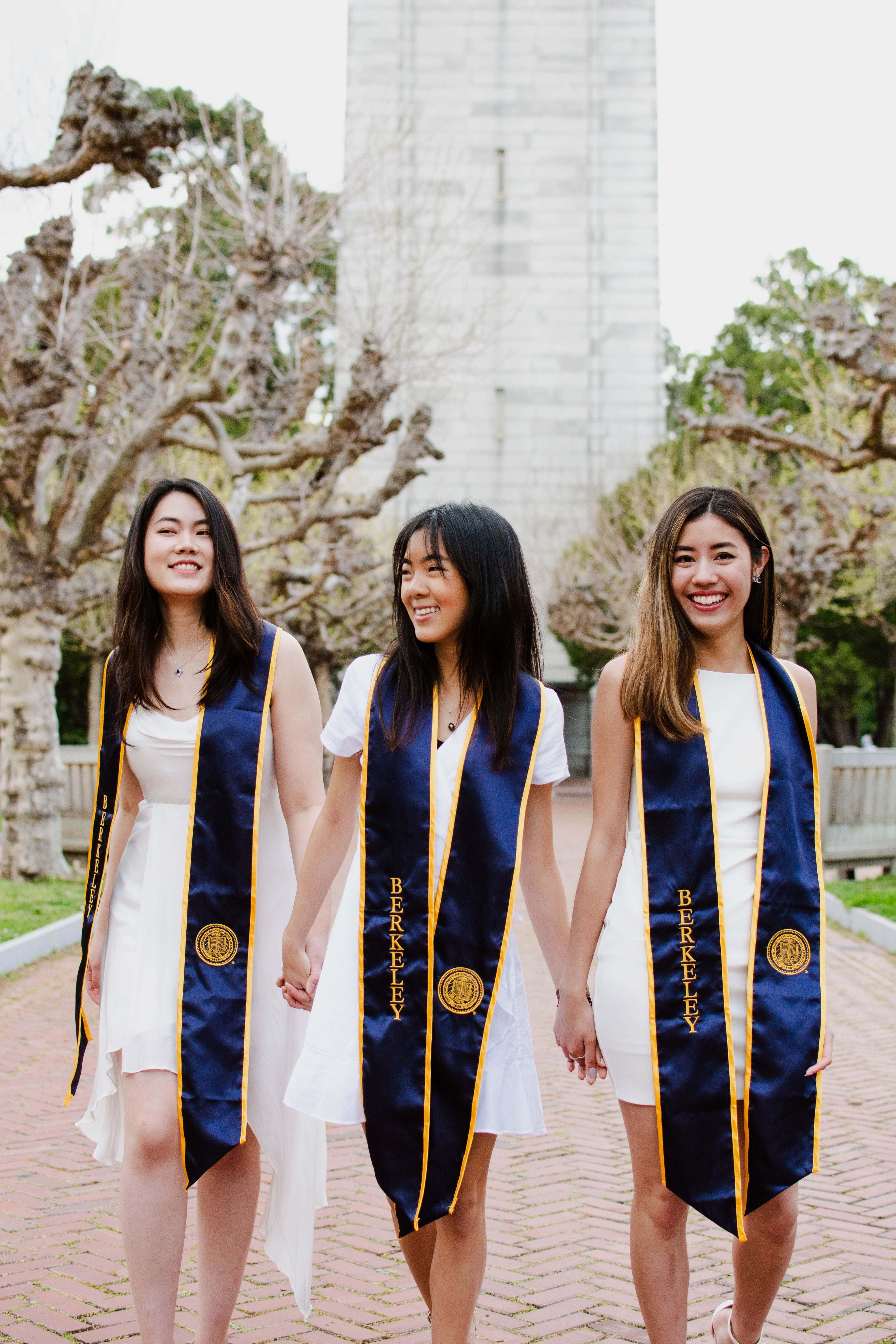 Graduation 桌布 壁紙 — 三位微笑的畢業生身穿藍金色柏克萊披肩，手牽手走在落葉樹下的磚路上。