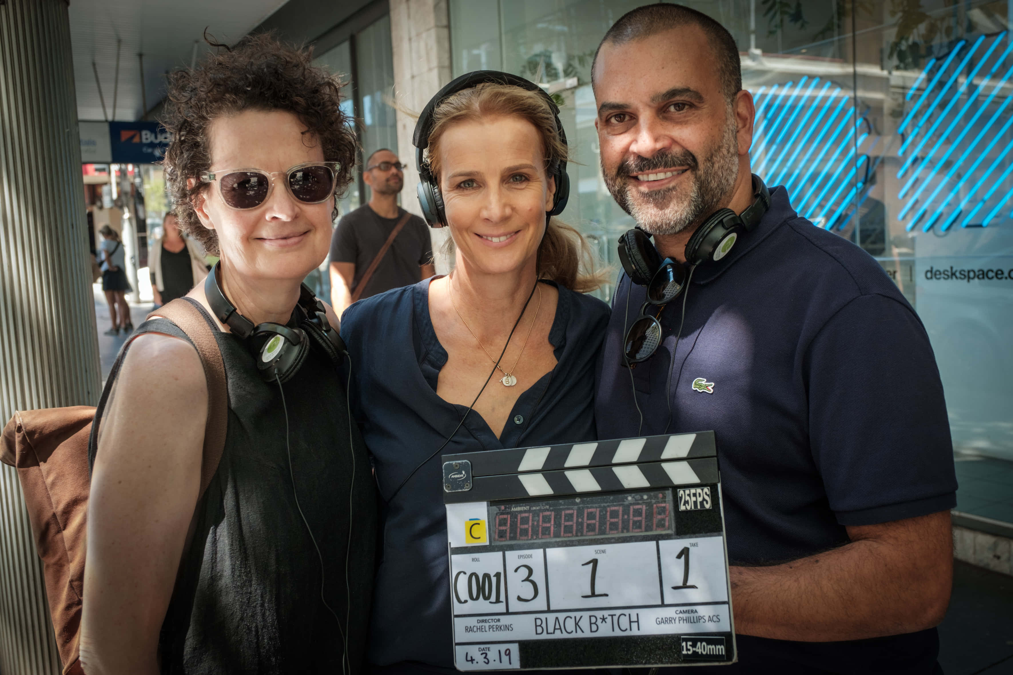 Rachel Griffiths 桌布 壁紙 — 三位微笑的電影工作人員在明亮的城市街道上拿著場記板擺姿勢。