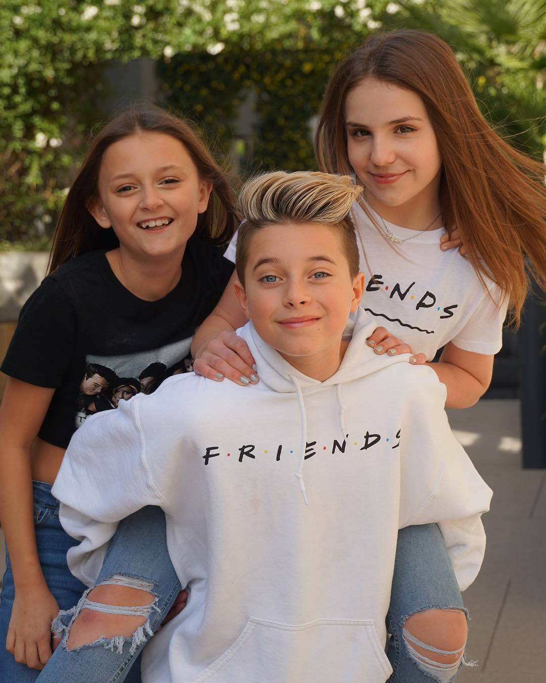 Piper Rockelle 桌布 壁紙 — 三位微笑的青少年穿著 Friends 主題連帽衫和破洞牛仔褲，在綠色樹葉前戶外擺姿勢。