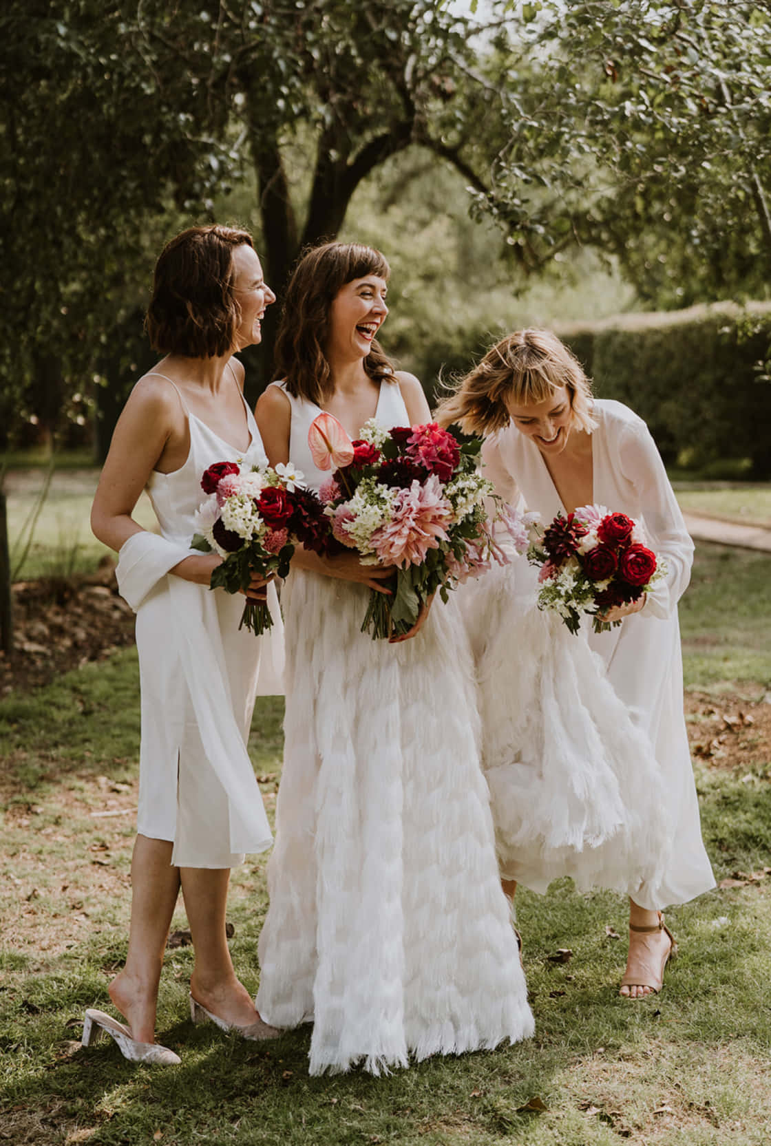 Bridal 桌布 壁紙 — 三位快樂的伴娘穿著白色羽毛裙，在陽光明媚的花園中手持鮮豔的紅色和粉紅色花束大笑。