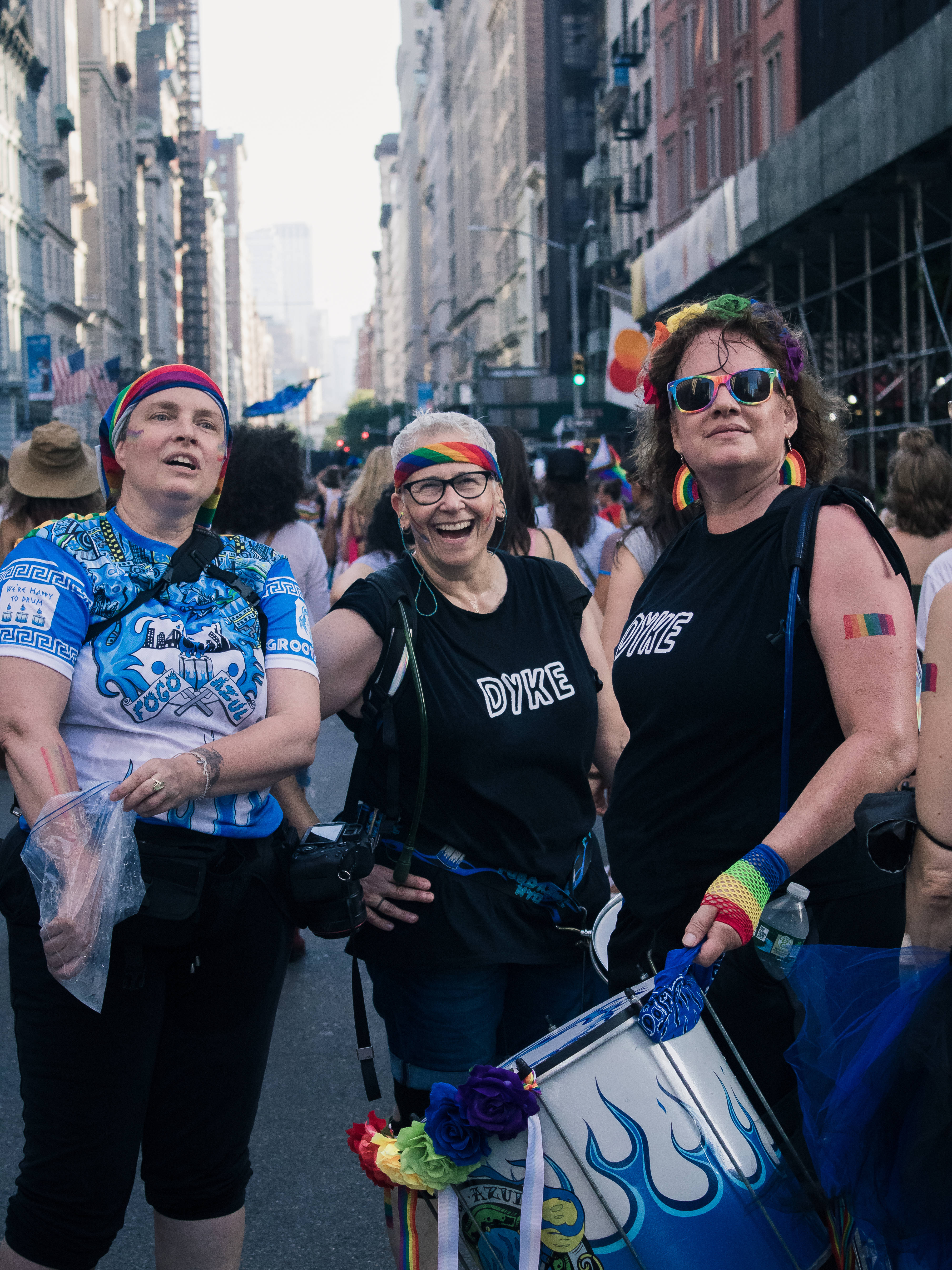 女同志 桌布 壁紙 — 三位快樂的女性，佩戴彩虹配飾，身穿「DYKE」T恤，在驕傲遊行期間穿過繁忙的城市街道。