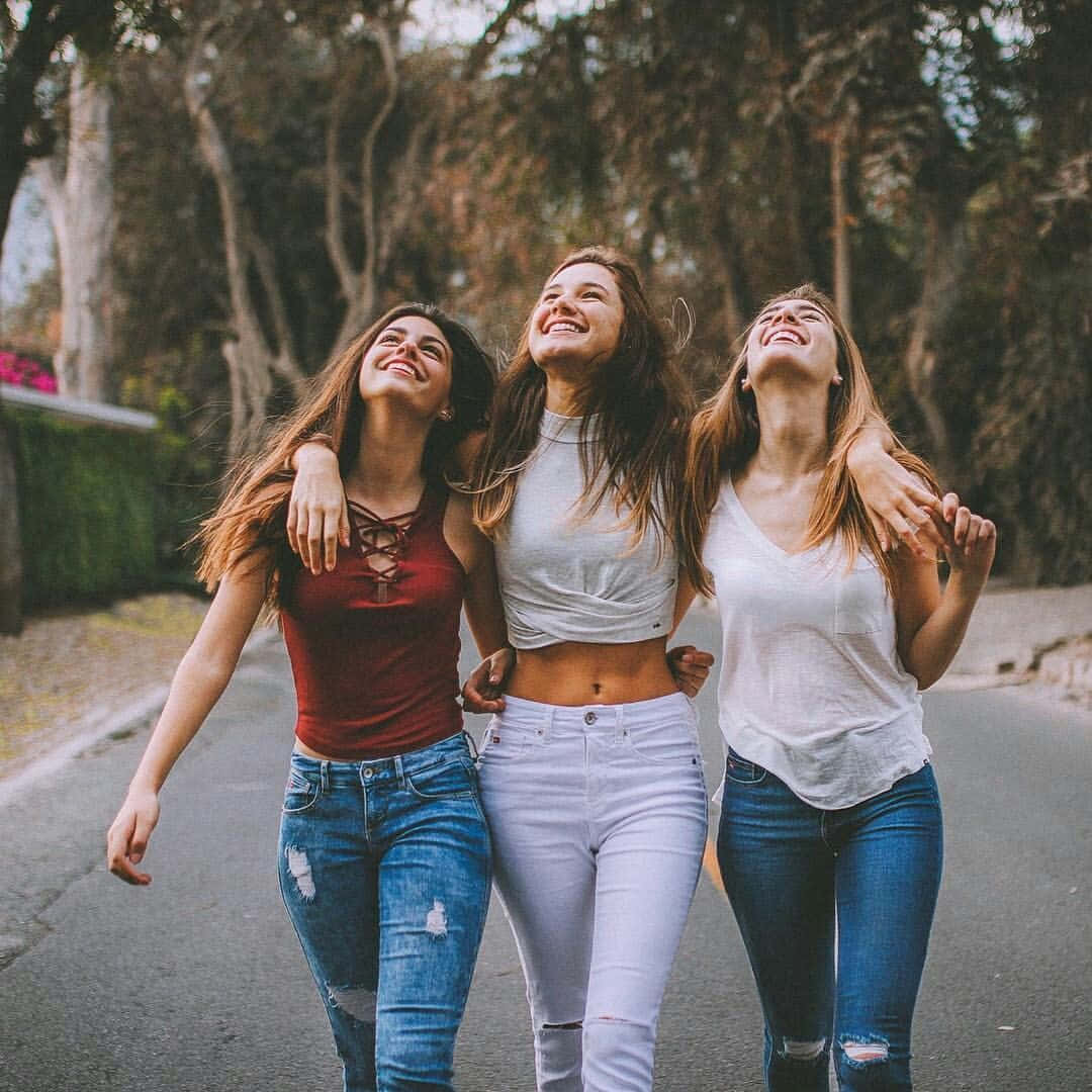 Friends 桌布 壁紙 — 三位快樂的年輕女性在陽光明媚的日子裡，笑著手挽手走在綠樹成蔭的街道上。