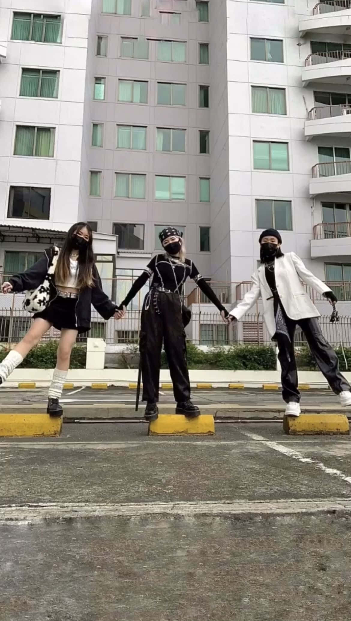 Friends 桌布 壁紙 — 三位戴著口罩的年輕女性穿著前衛街頭服飾，在現代公寓大樓前的黃色停車塊上擺出同步姿勢。
