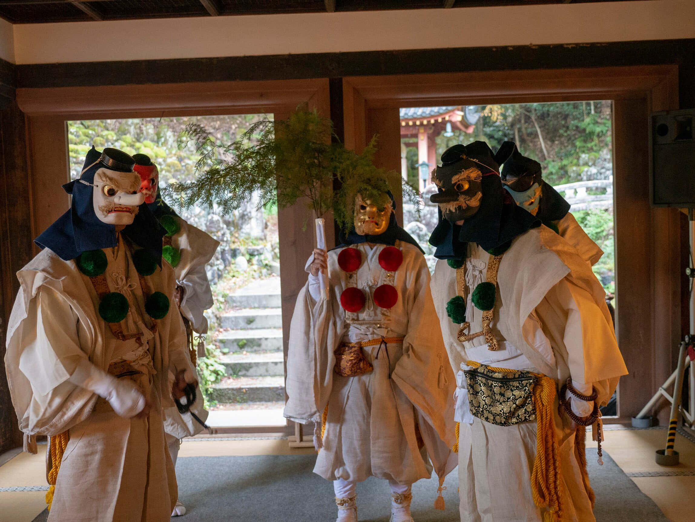 Tengu 桌布 壁紙 — 三位戴面具的日本表演者穿著傳統儀式服裝，站在木造神社內，外面可見綠意。