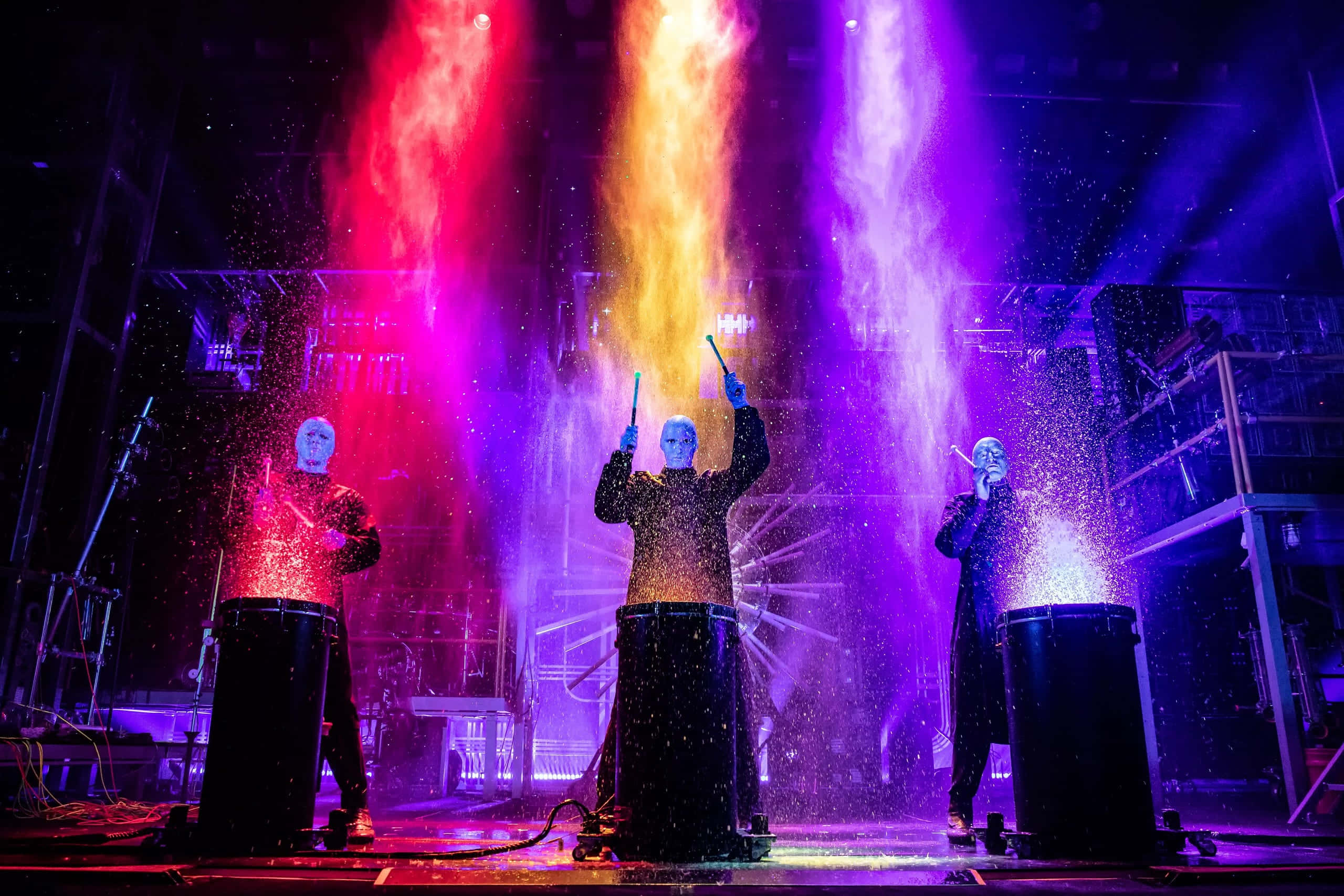 Blue Man Group 桌布 壁紙 — 三位戴面具的表演者在鮮豔的紅、紫、黃燈光下打鼓，閃爍的火花飛舞。